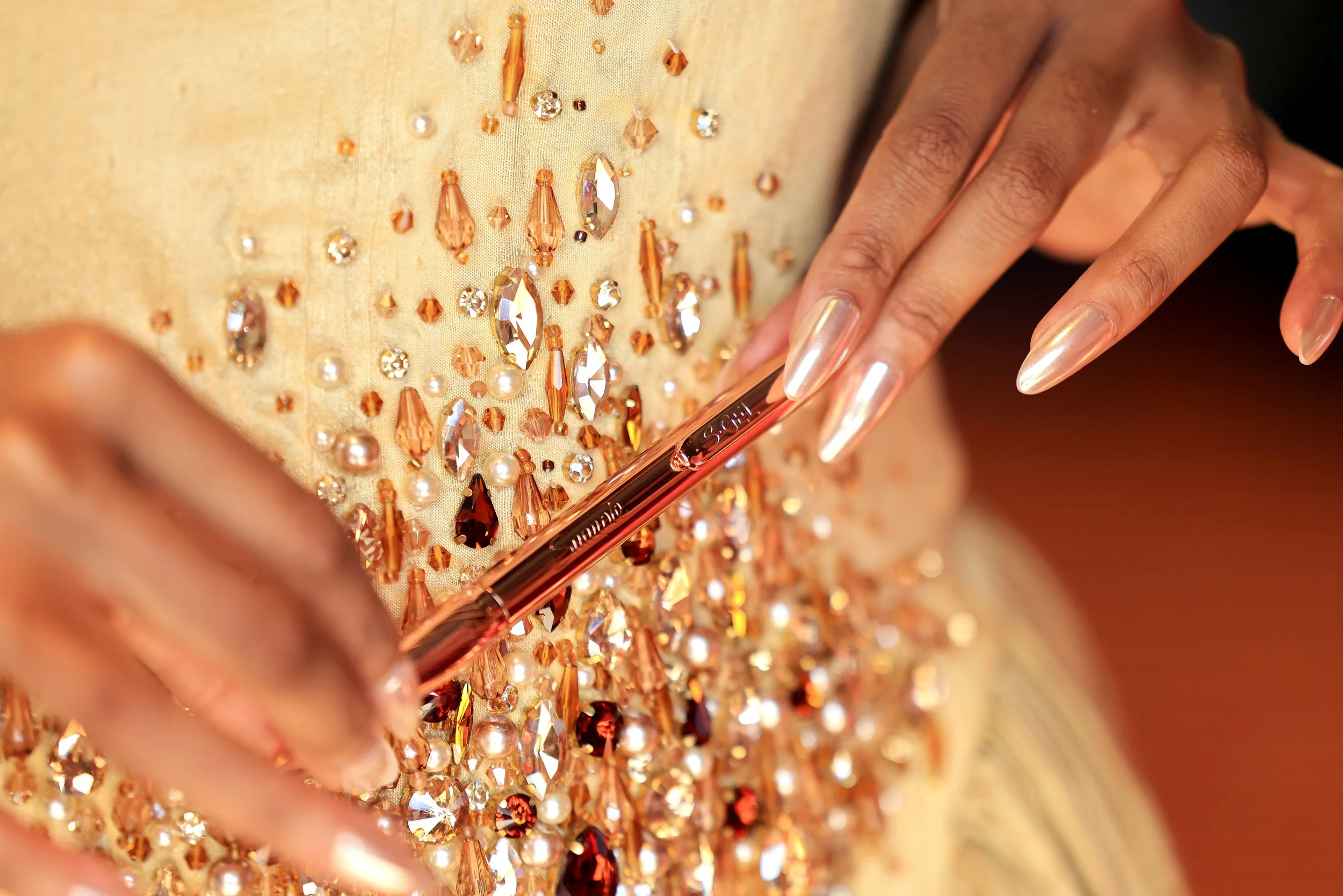 Close-up of a person's hand holding a rose gold makeup brush, resting on a richly embellished fabric adorned with various crystals, beads, and pearls in shades of gold, orange, and white.