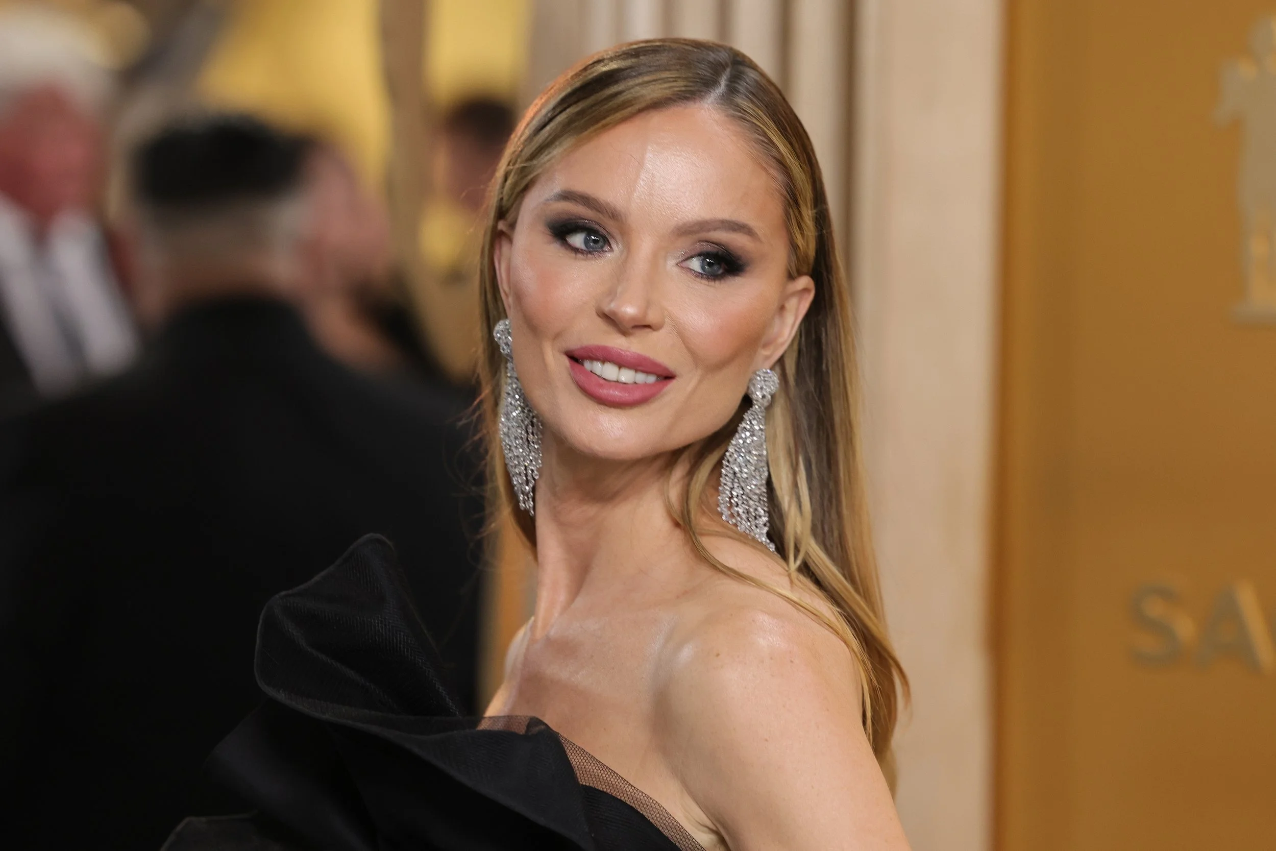 A woman with long blonde hair and blue eyes wearing large, dangling, sparkly earrings and a black dress with ruffled shoulder detail, attending a formal event.