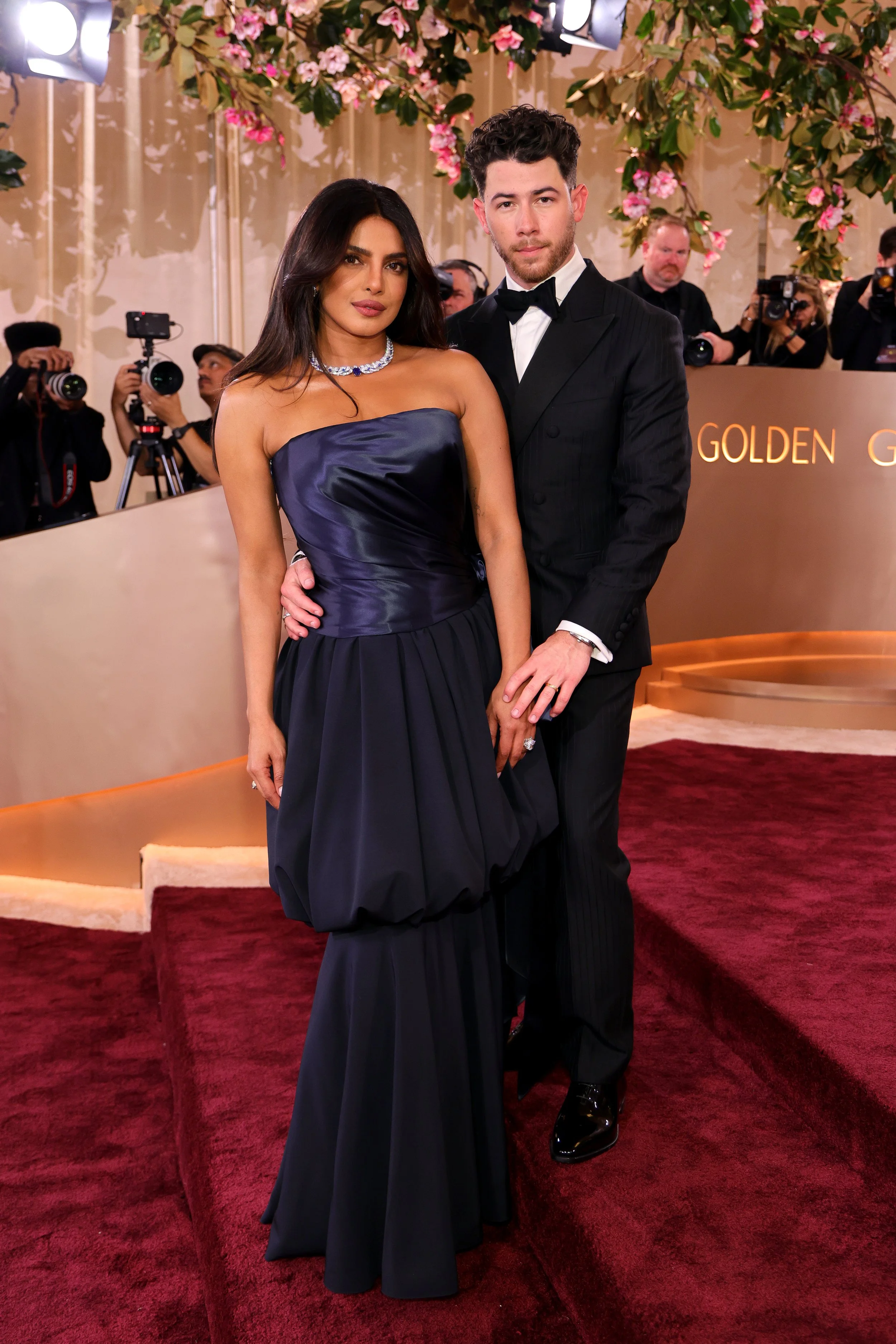 A woman in a strapless navy gown and a man in a black tuxedo pose together on the red carpet at a glamorous event, with photographers in the background and floral decorations overhead.