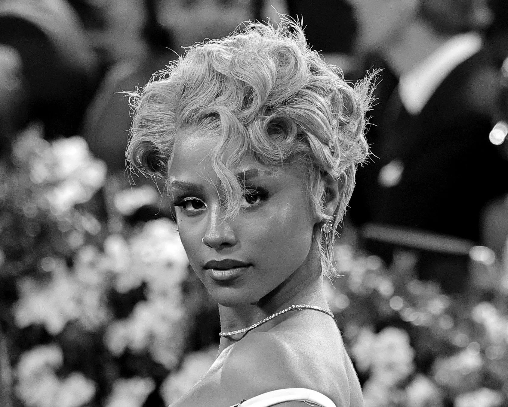 A woman with curly, platinum blonde hair styled in vintage waves, wearing earrings and a necklace, looking directly at the camera with a confident expression, blurred floral background.