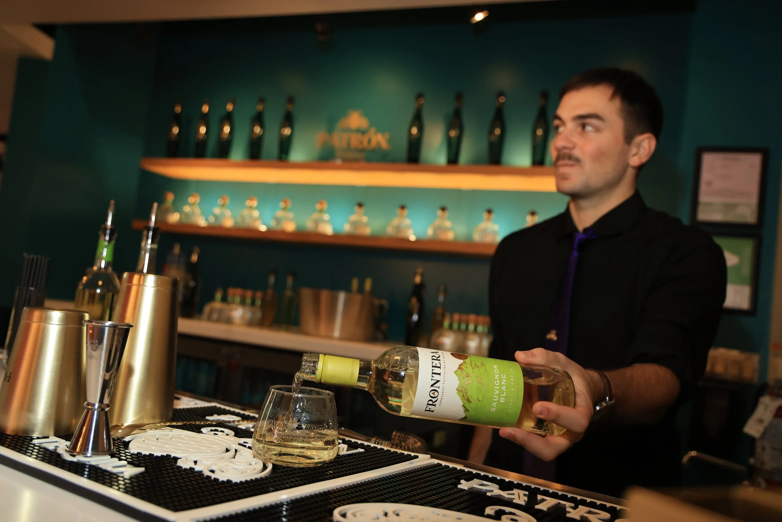 A bartender pours white wine into a glass at a bar counter with various bar tools and bottles, with shelves holding decorative bottles and awards in the background.
