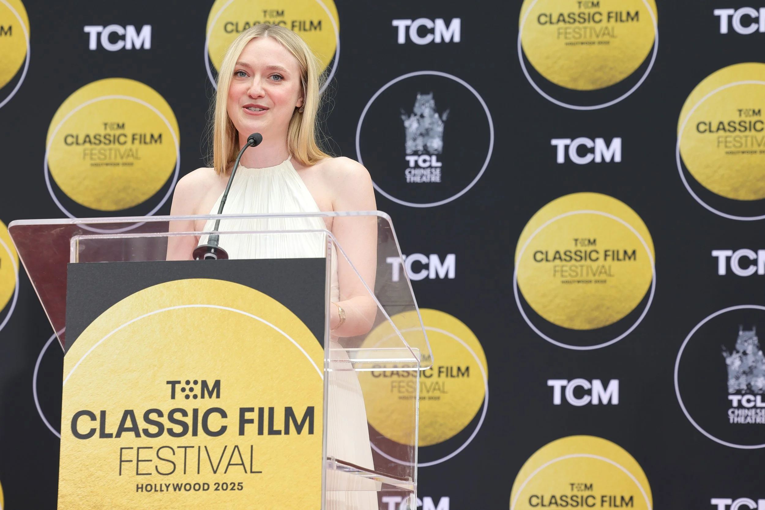 A woman with blonde hair speaking at a podium during the TCM Classic Film Festival in Hollywood 2025. The backdrop features multiple TCM and TCL Chinese Theatre logos.