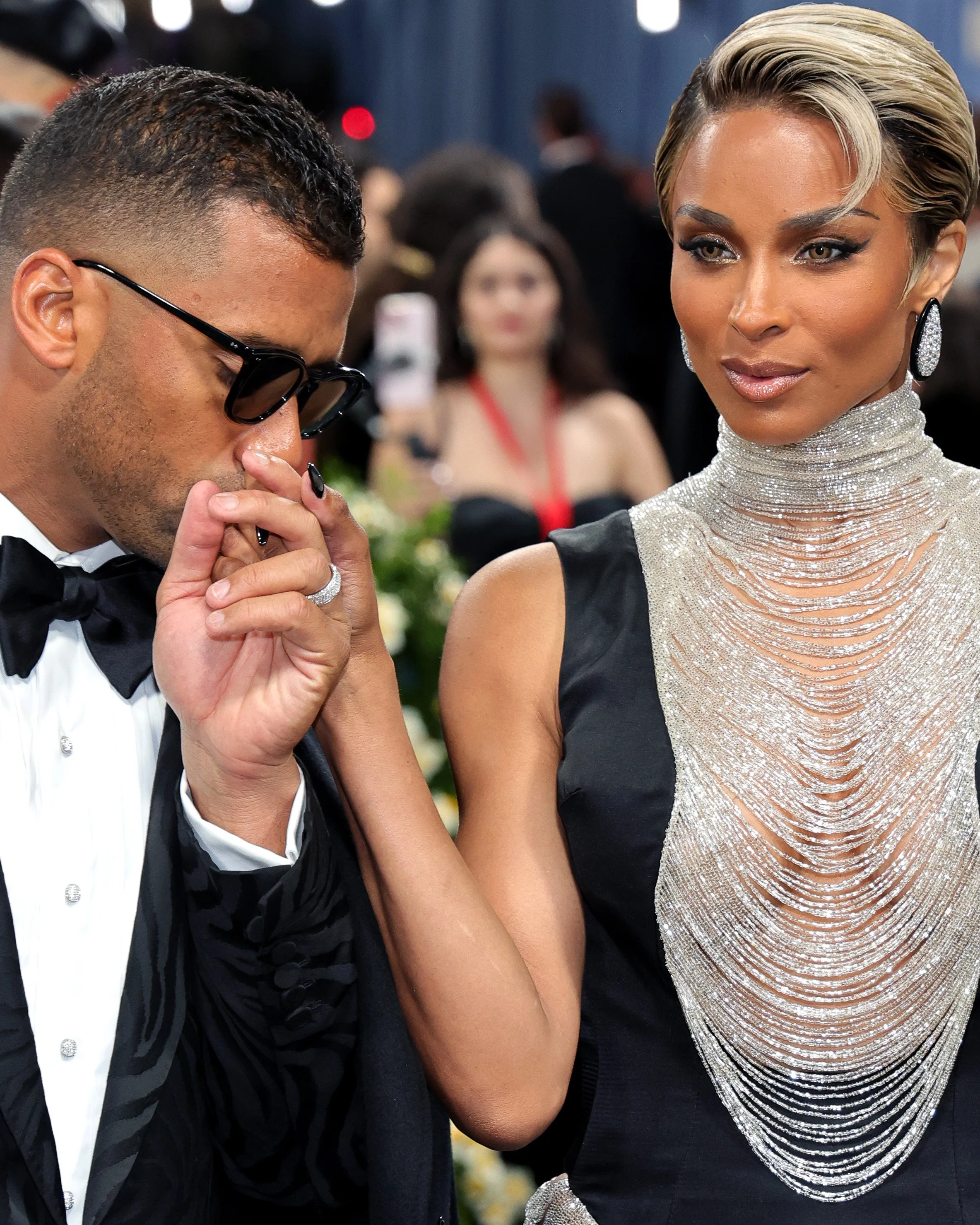 A man dressed in a tuxedo kisses the hand of a woman wearing a glamorous black and silver dress with a high neckline, at a formal event.