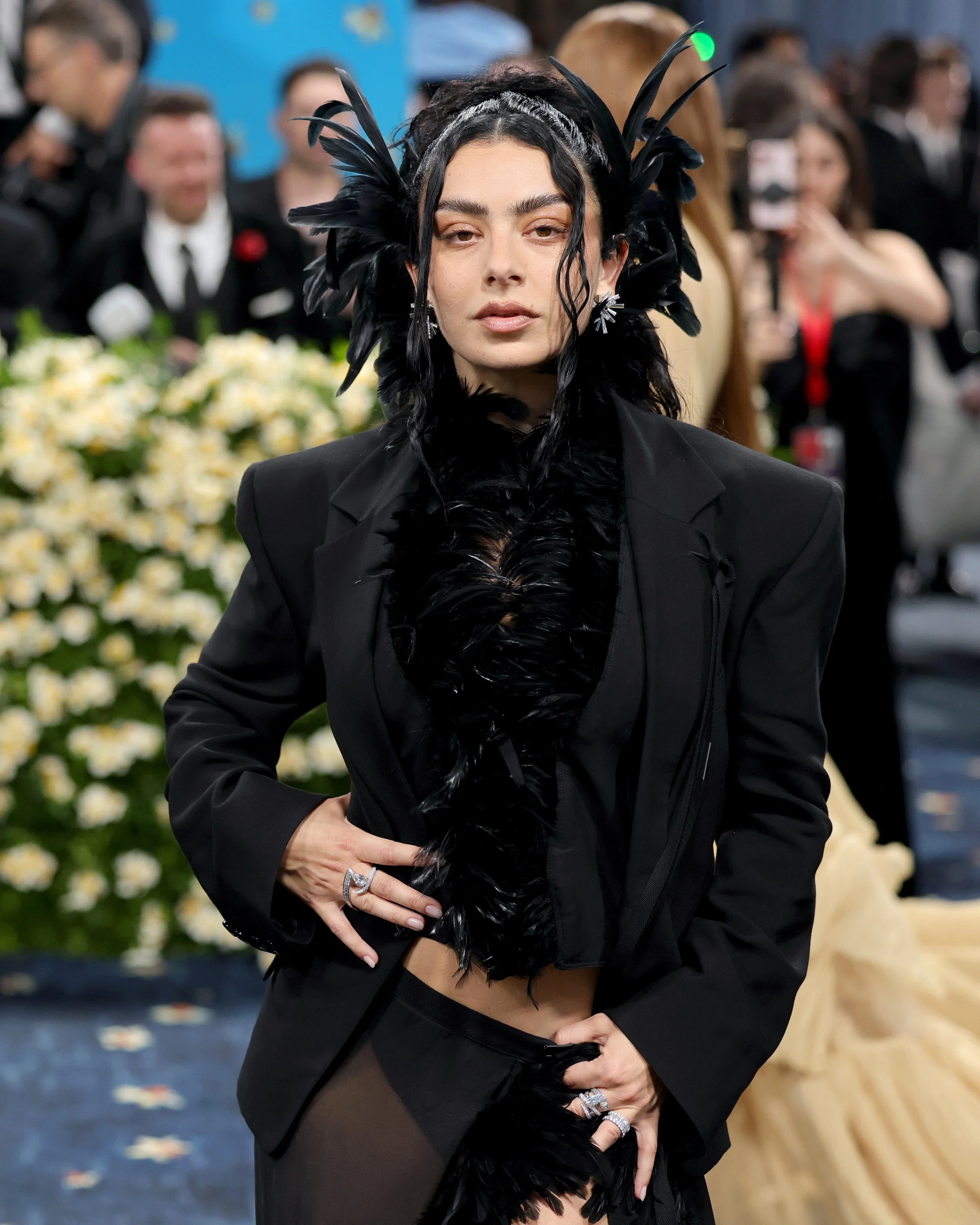 A woman at a fashion event wearing a black feathered headpiece and a black blazer with feathered details, standing in front of a floral backdrop with photographers and attendees in the background.