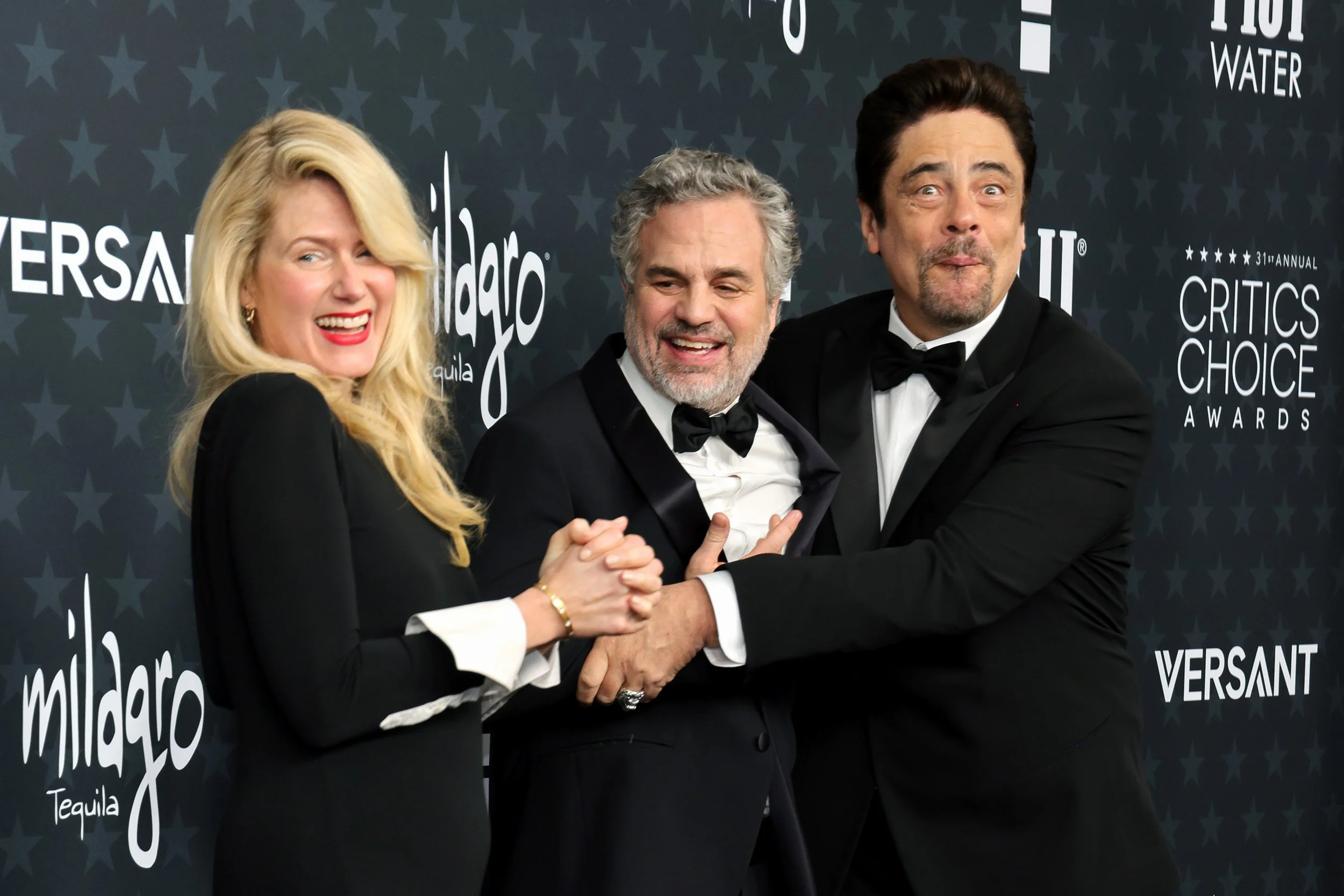 Three people, two men and one woman, dressed in black tuxedos, holding hands and embracing each other on the red carpet at an awards event, with a black backdrop featuring logos and text.