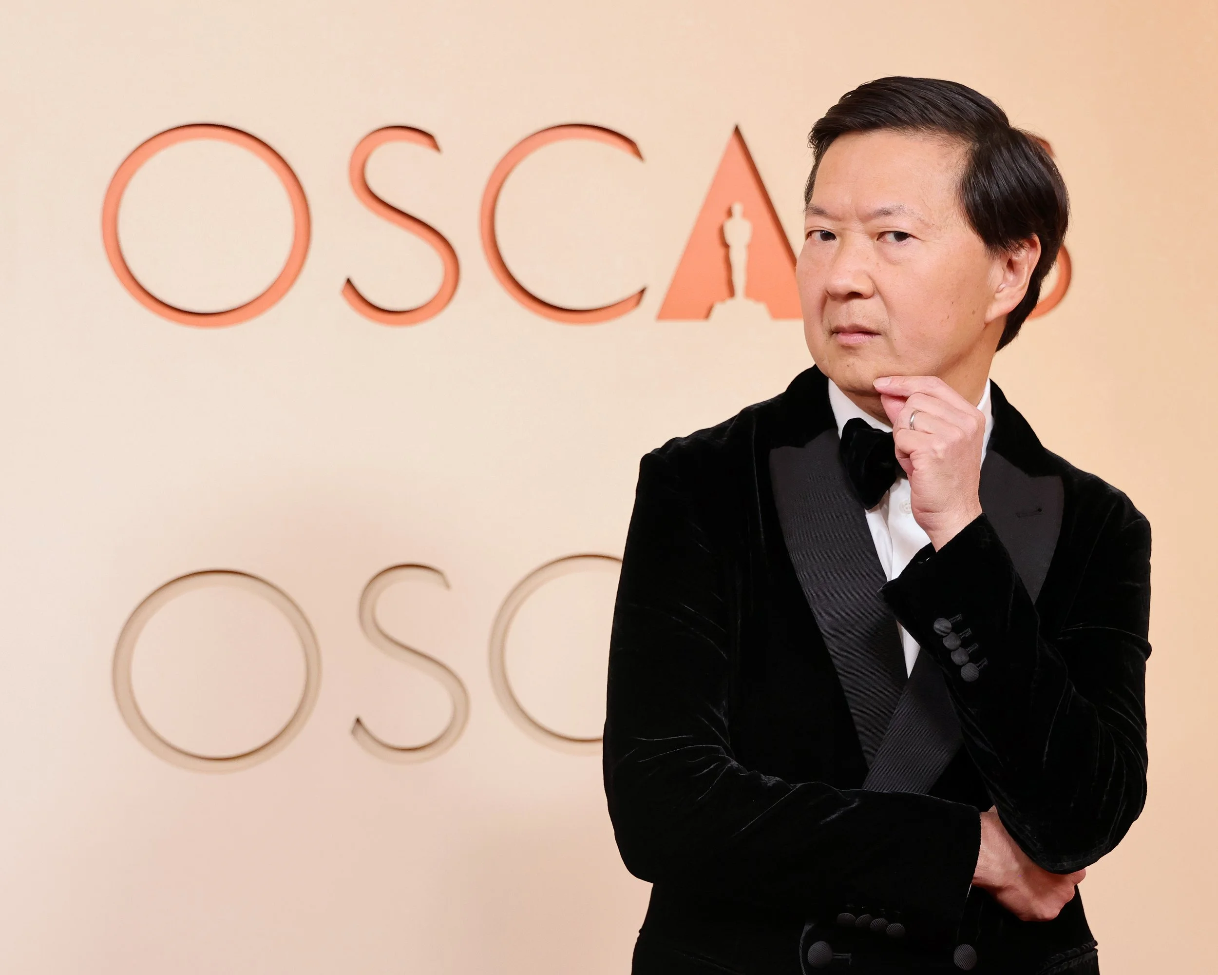 Person in tuxedo standing in front of a wall with the words 'Oscar' written on it, holding chin and looking thought-provoking.
