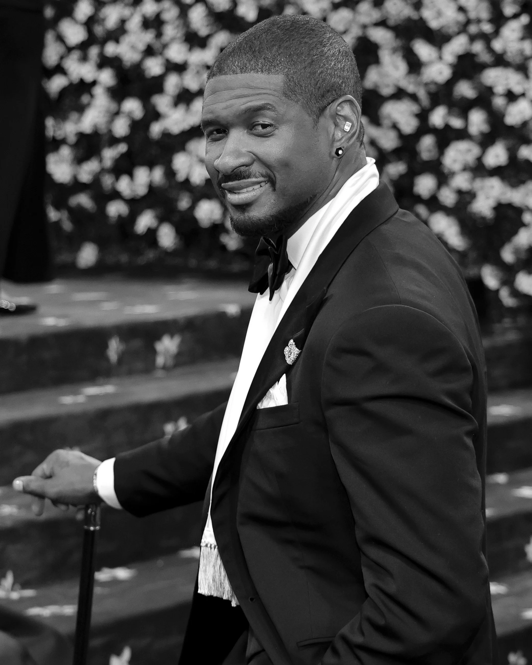A man in a tuxedo with a bow tie, earrings, and a press pin, smiling and looking over his shoulder at the camera, holding a cane in one hand, standing in front of a floral backdrop.