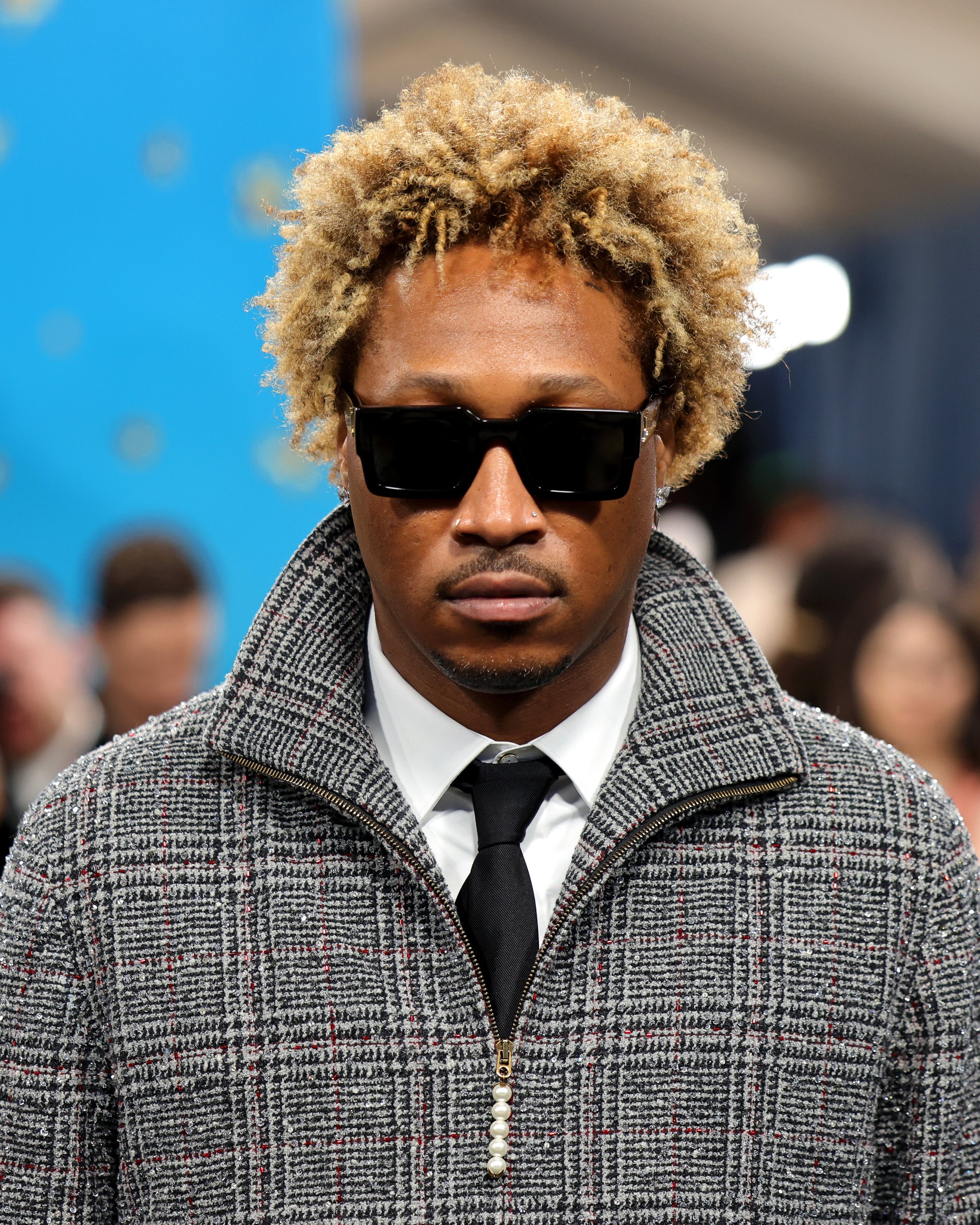 A man with blonde curly hair wearing black sunglasses, a checked coat, white shirt, and black tie at a crowded indoor event.