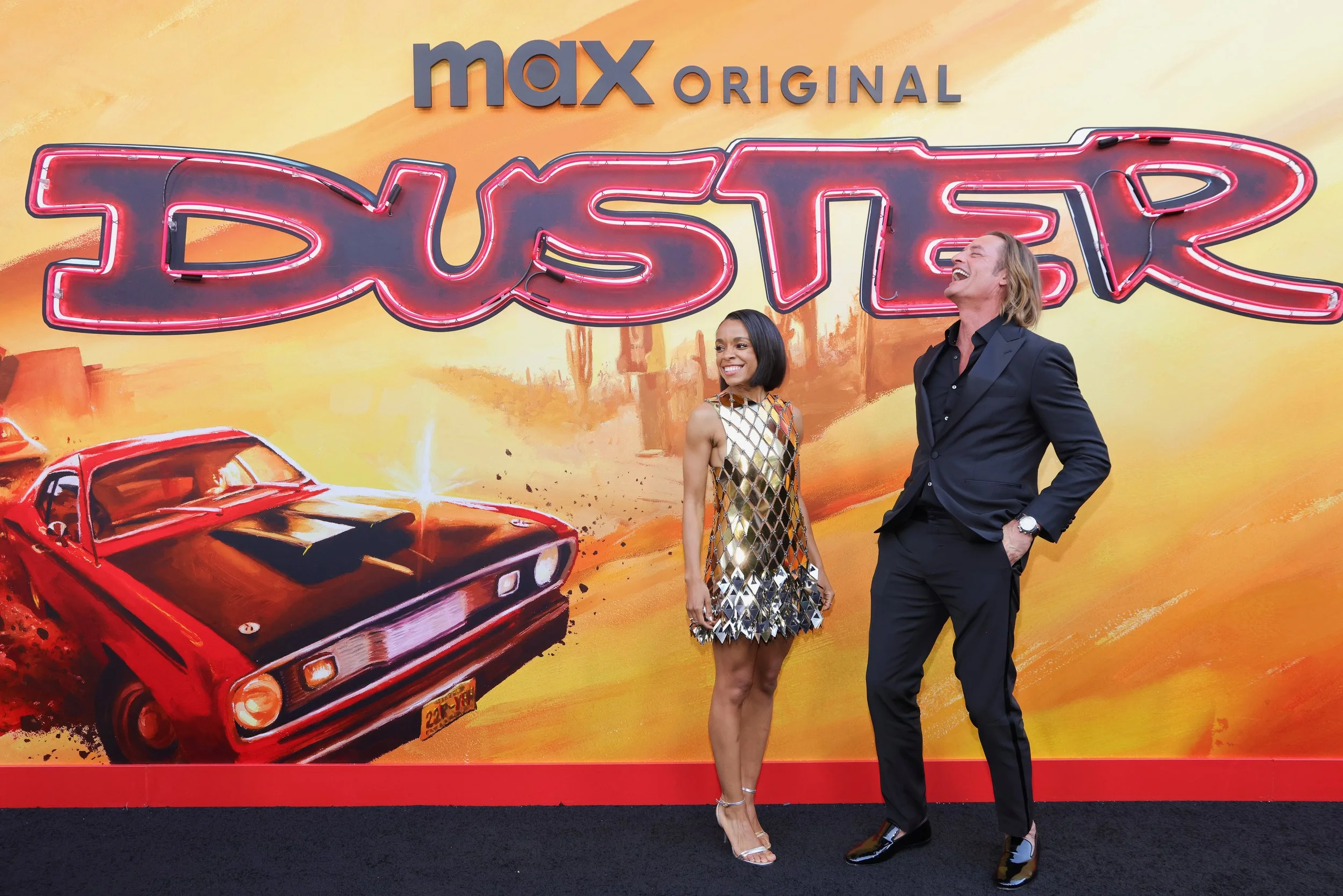 Two people smiling and standing in front of a promotional backdrop for the movie 'Dustup.' The backdrop features a red muscle car and a desert scene with cacti and a sunset. The woman is wearing a metallic, diamond-patterned dress and high heels, whi