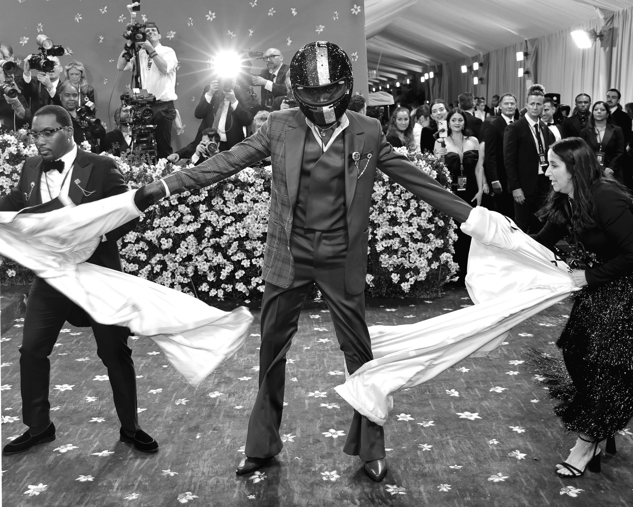 Person wearing a helmet and suit dancing with two women holding long fabric strips at a lively event with photographers and audience in the background.