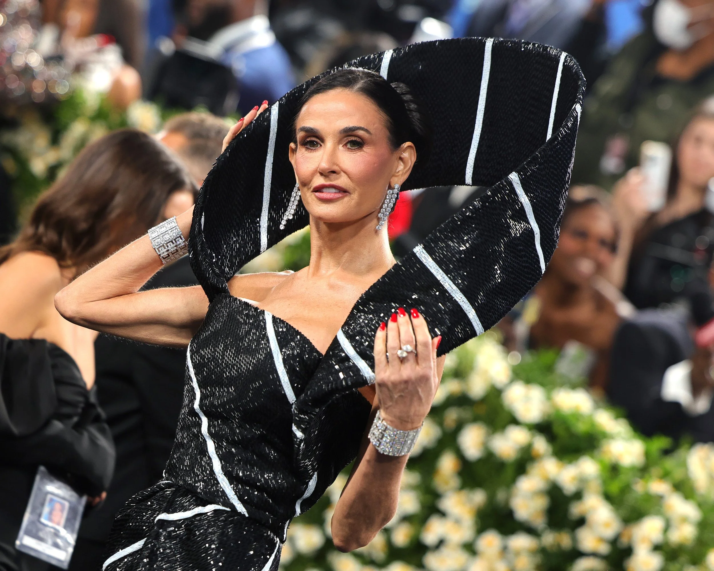 Woman in a black and white sequined dress with large matching hat, jewelry, and red nail polish, attending a formal event with a floral arrangement and people in the background.