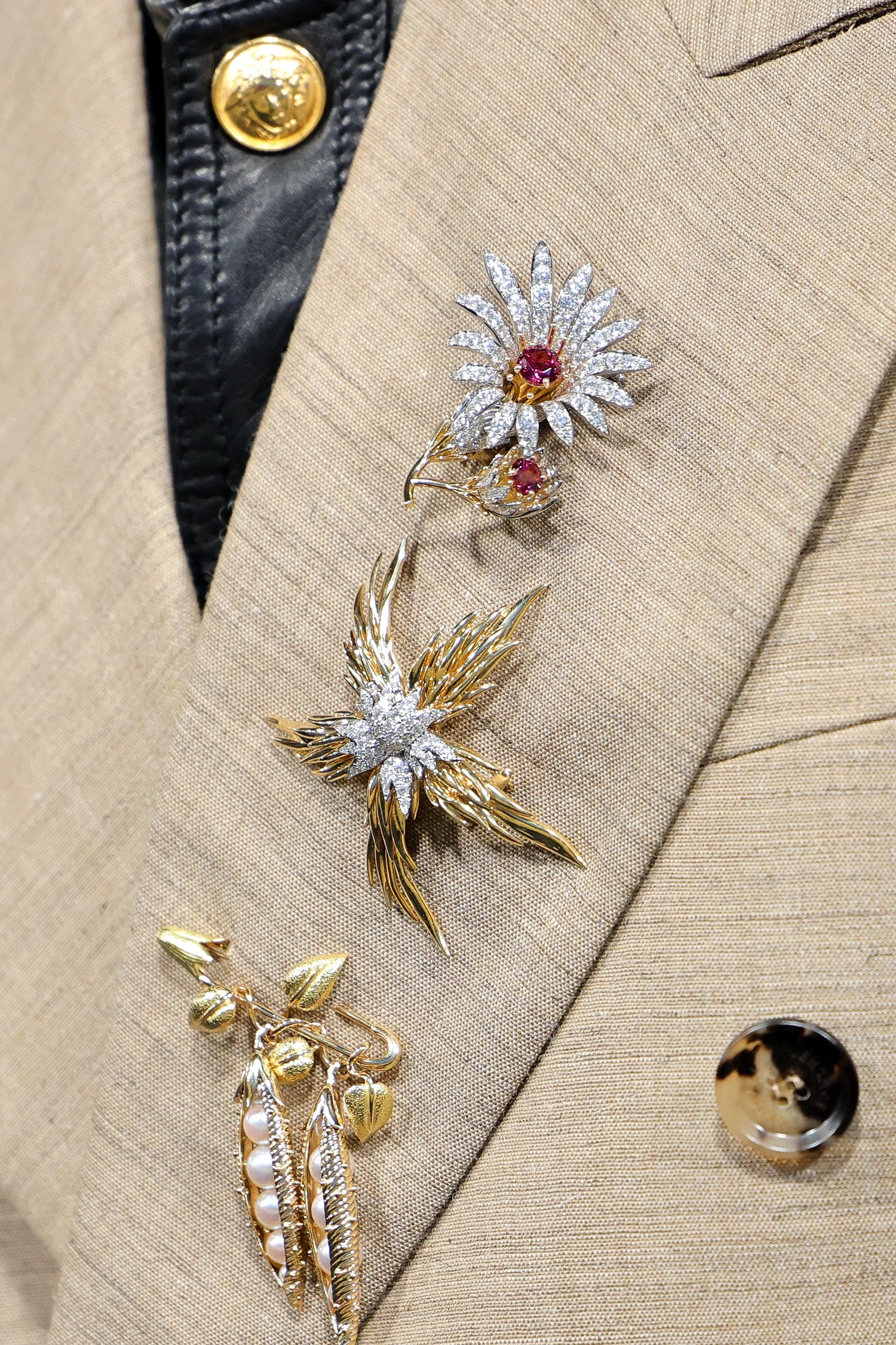 A beige suit jacket with gold buttons and decorative pins: a flower-shaped pin with red and white stones, a bird pin with gold and white stones, and a leaf and pearl pin.