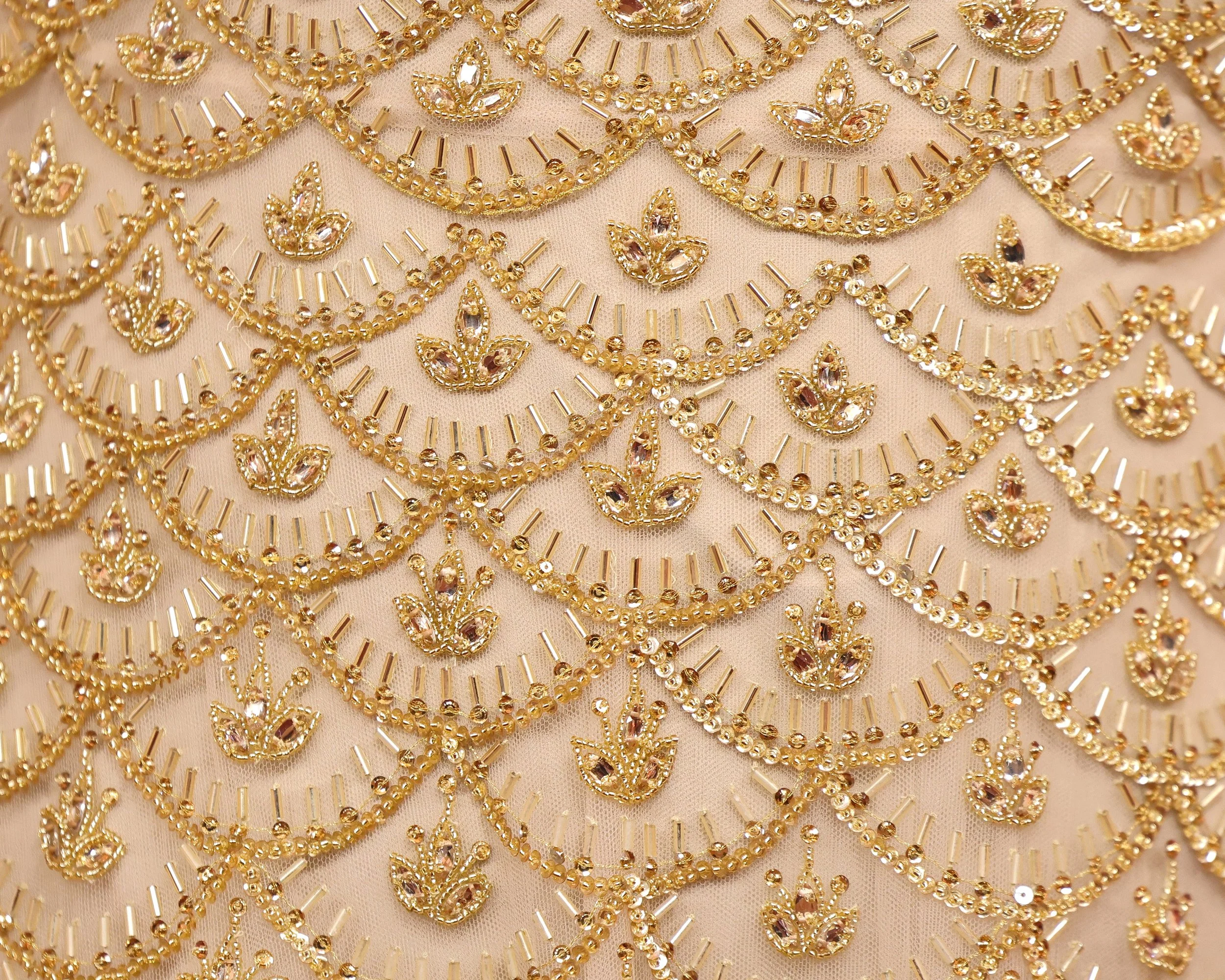 Close-up view of intricate gold embroidery with small crystals on a pink fabric background, arranged in a scalloped pattern.