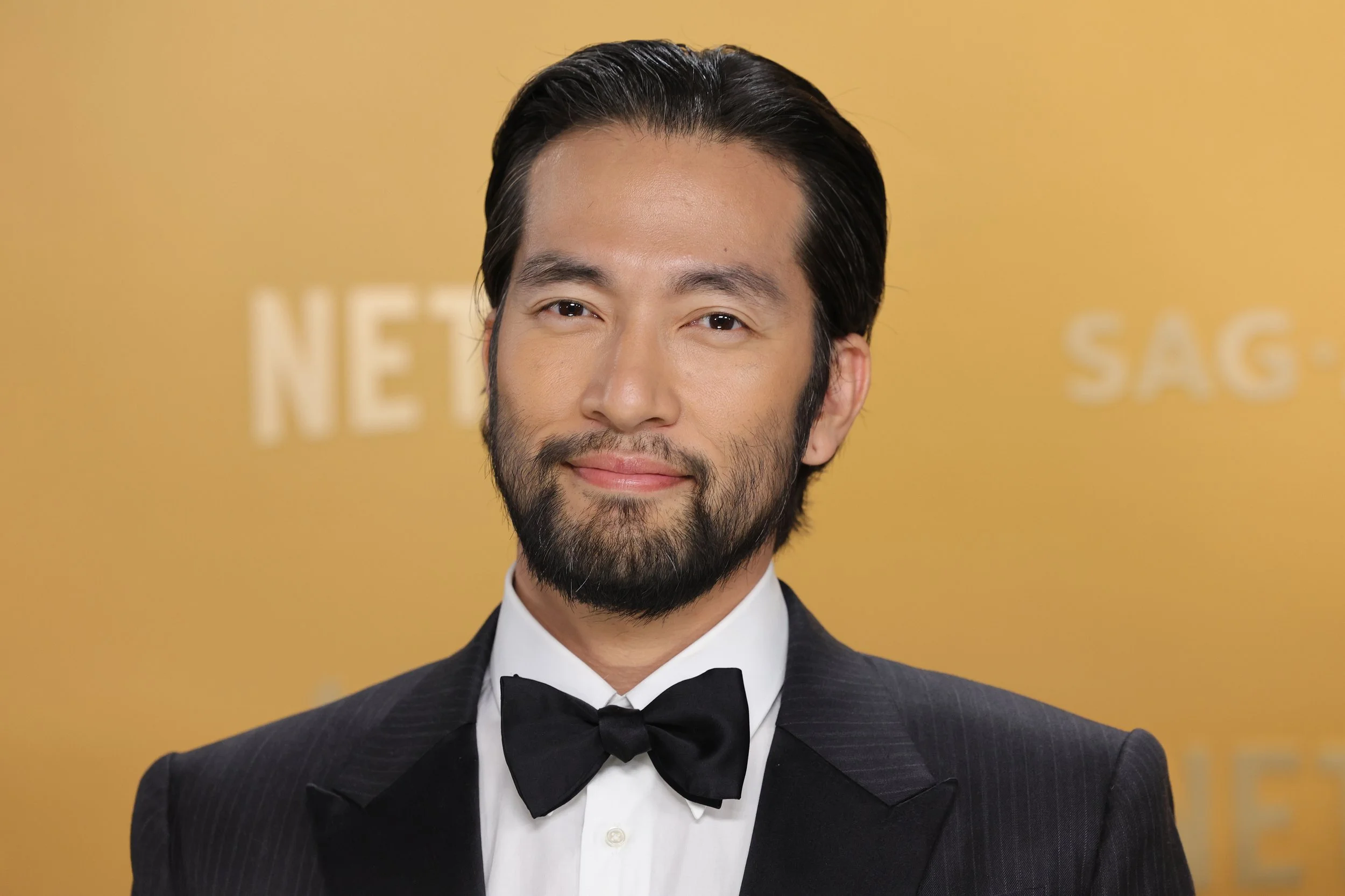 A man with a beard and slicked-back dark hair, dressed in a black tuxedo with a white shirt and black bow tie, smiling at the camera.