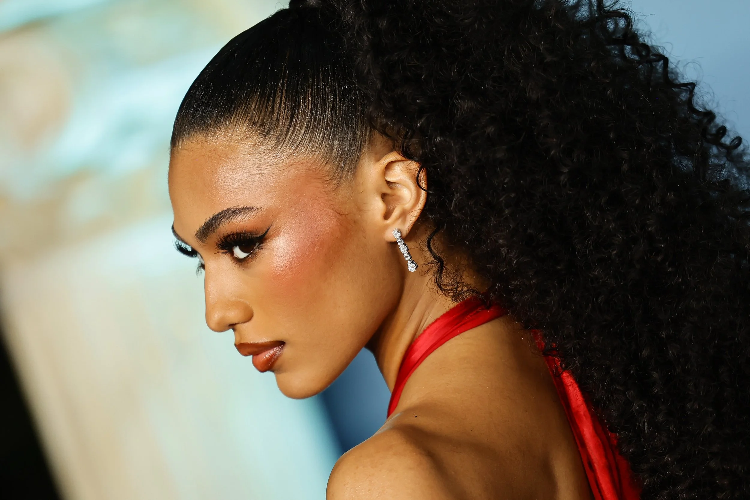 A woman with dark, curly hair styled in a high ponytail, wearing diamond earrings and a red dress.
