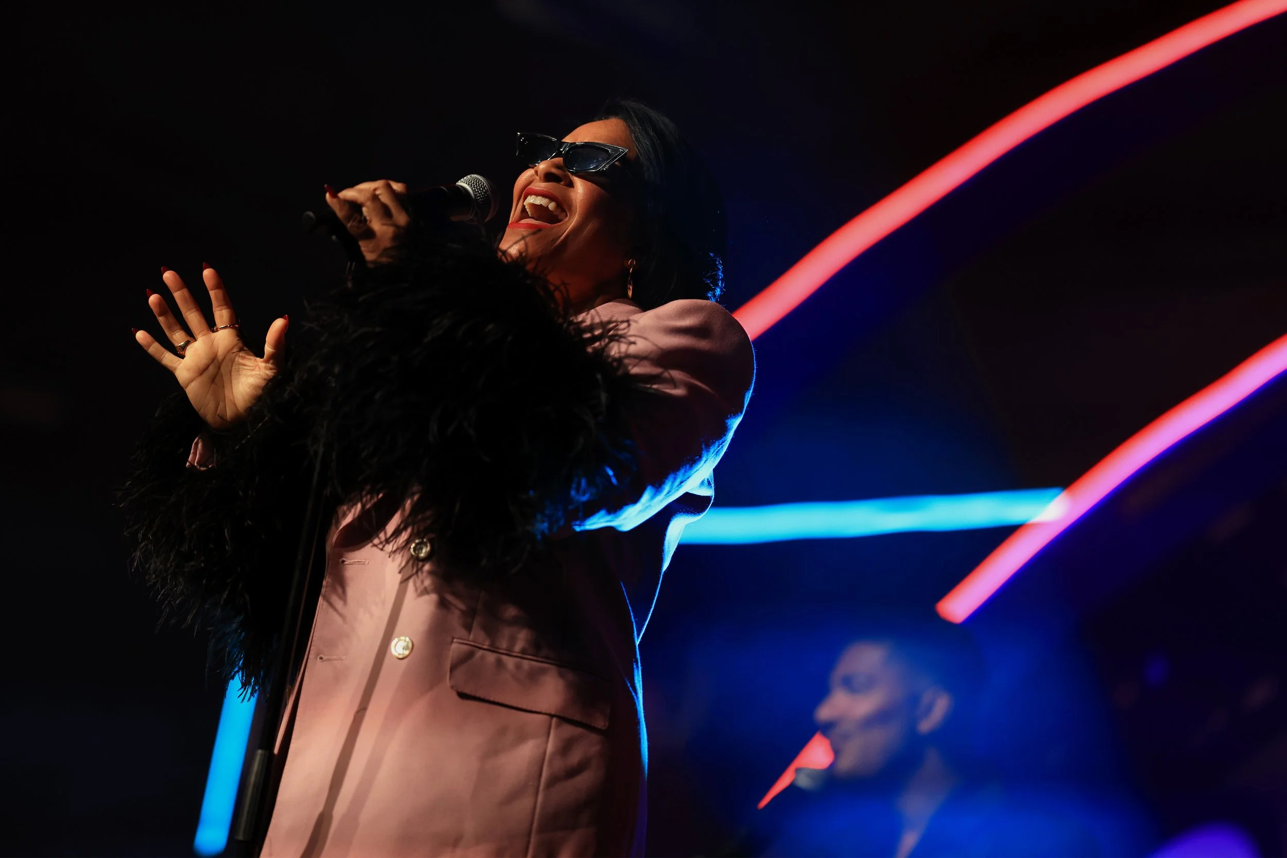 A woman singing on stage, wearing sunglasses and a pink blazer with black feathered sleeves, illuminated by colorful stage lights.