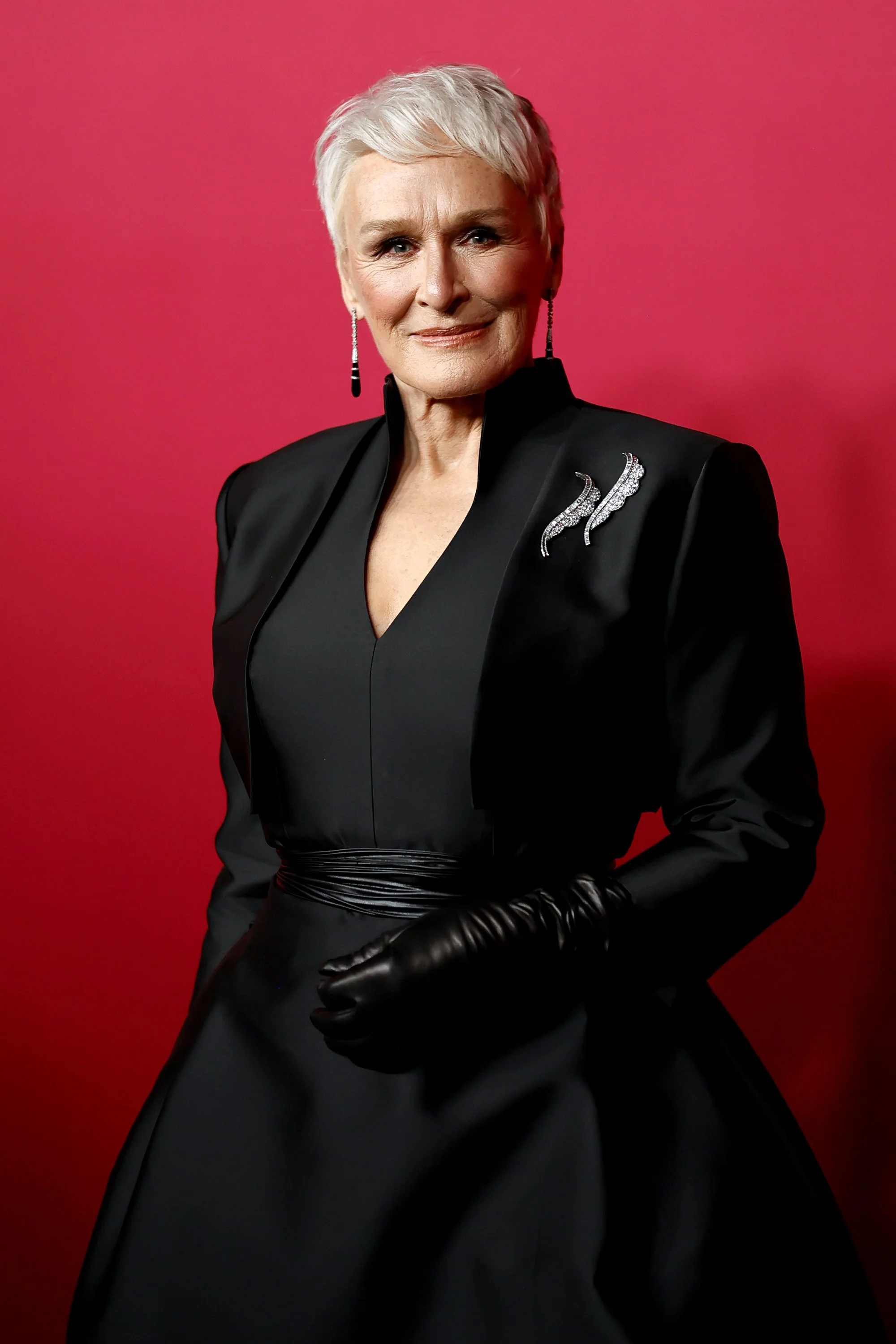 An elegant older woman with short, white hair, wearing a black jacket with shoulder embellishments, a black dress, long black gloves, and dangling earrings, standing against a red background.