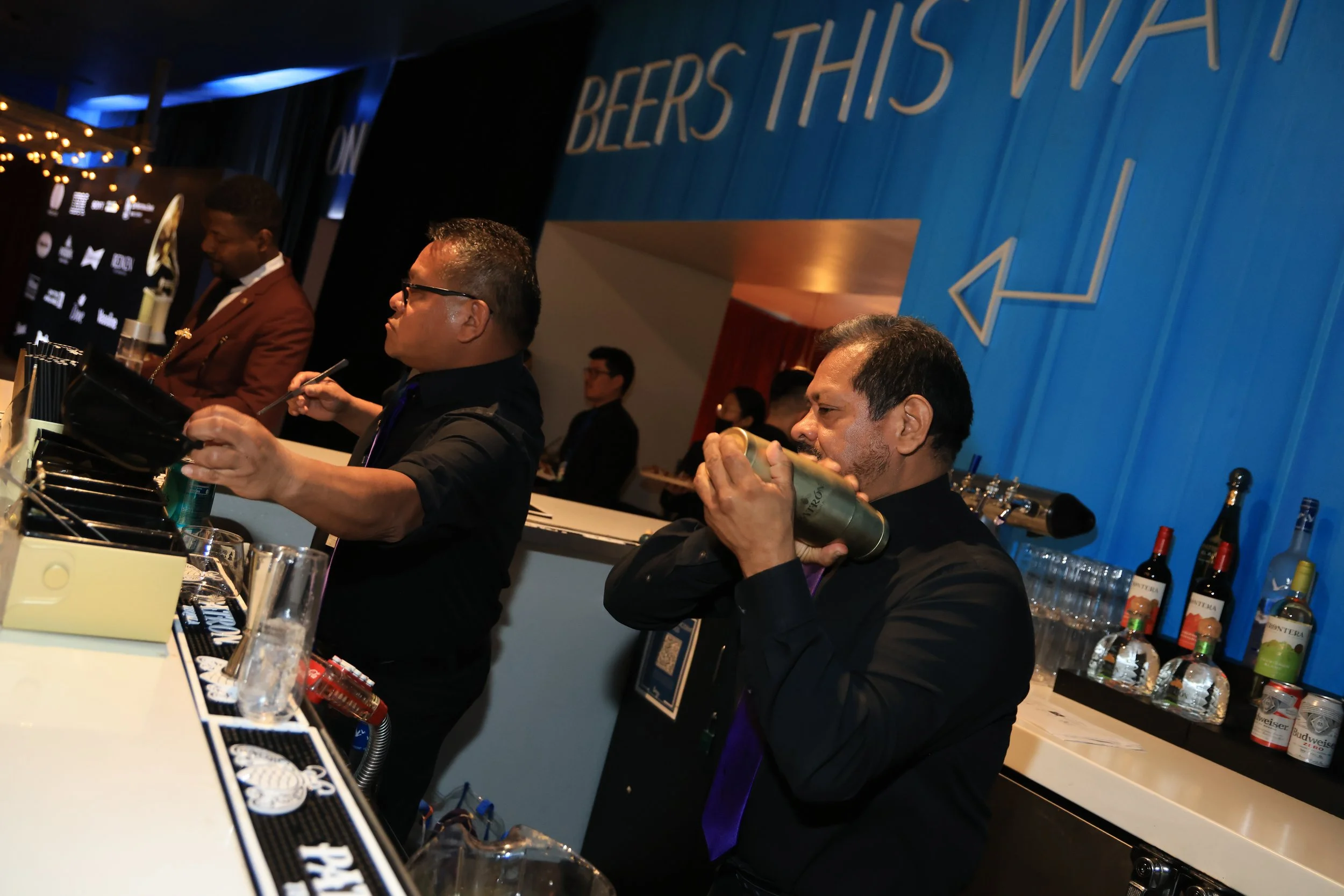 Bartenders preparing drinks at a bar, with one shaking a cocktail shaker and another pouring liquor into a glass, in front of a blue wall with a sign pointing left that says 'BEERS THIS WAY.'