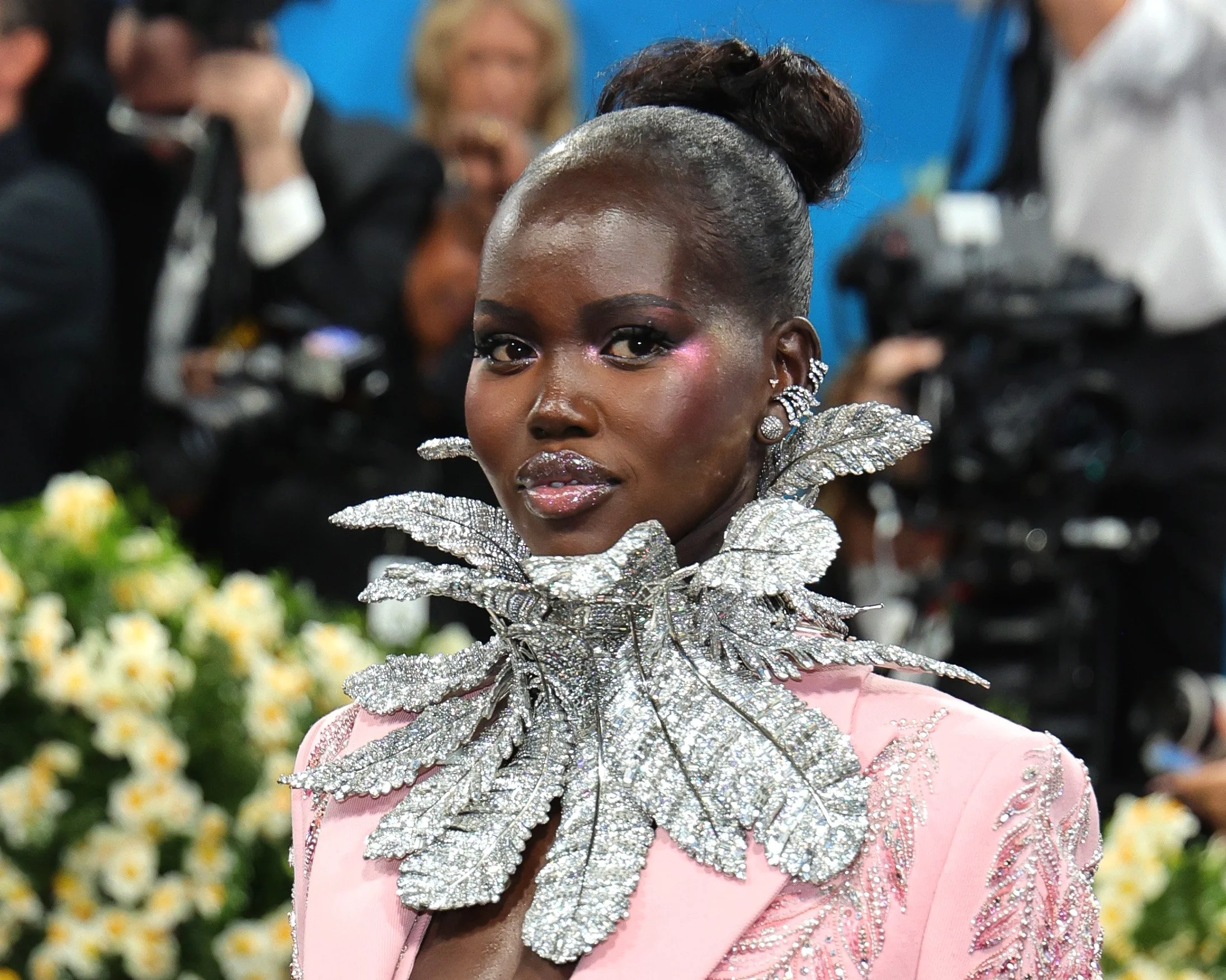 A woman wearing a pink dress with intricate embroidery and a large, shimmering leaf-shaped necklace, at a formal event with a blurred background of people and cameras.