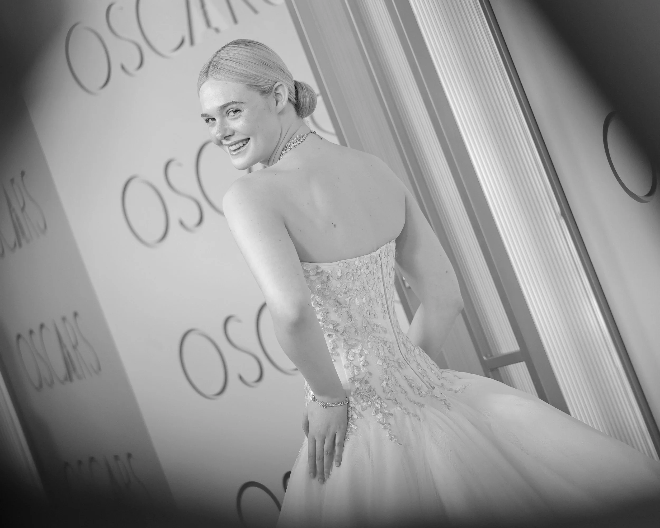 A woman in a wedding dress smiling and looking over her shoulder at the camera, standing in front of a backdrop with the word 'OSCAR' printed repeatedly.