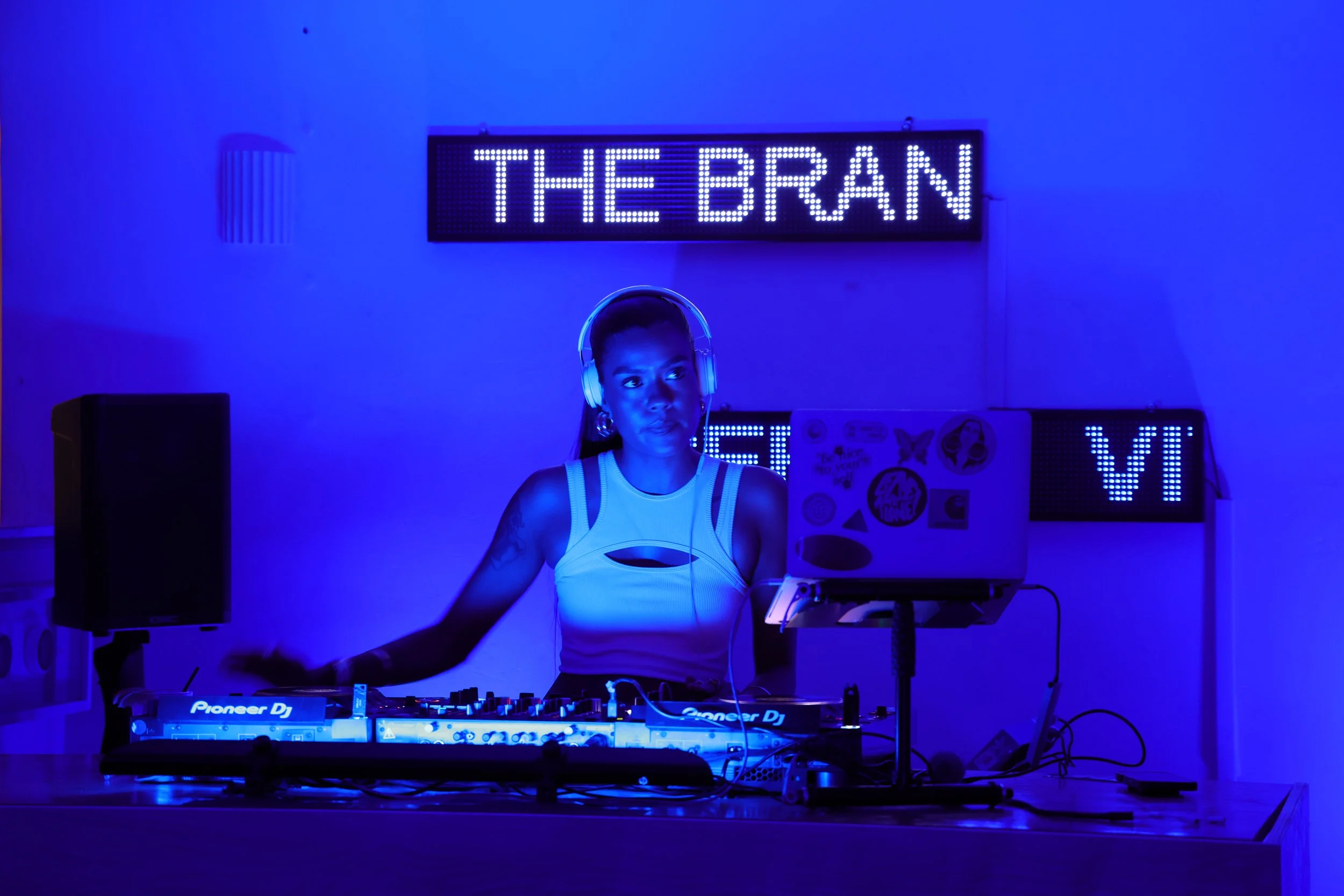A female DJ wearing headphones behind a turntable in a dimly lit room illuminated by blue LED lights, with a sign reading 'THE BRAN' in the background.
