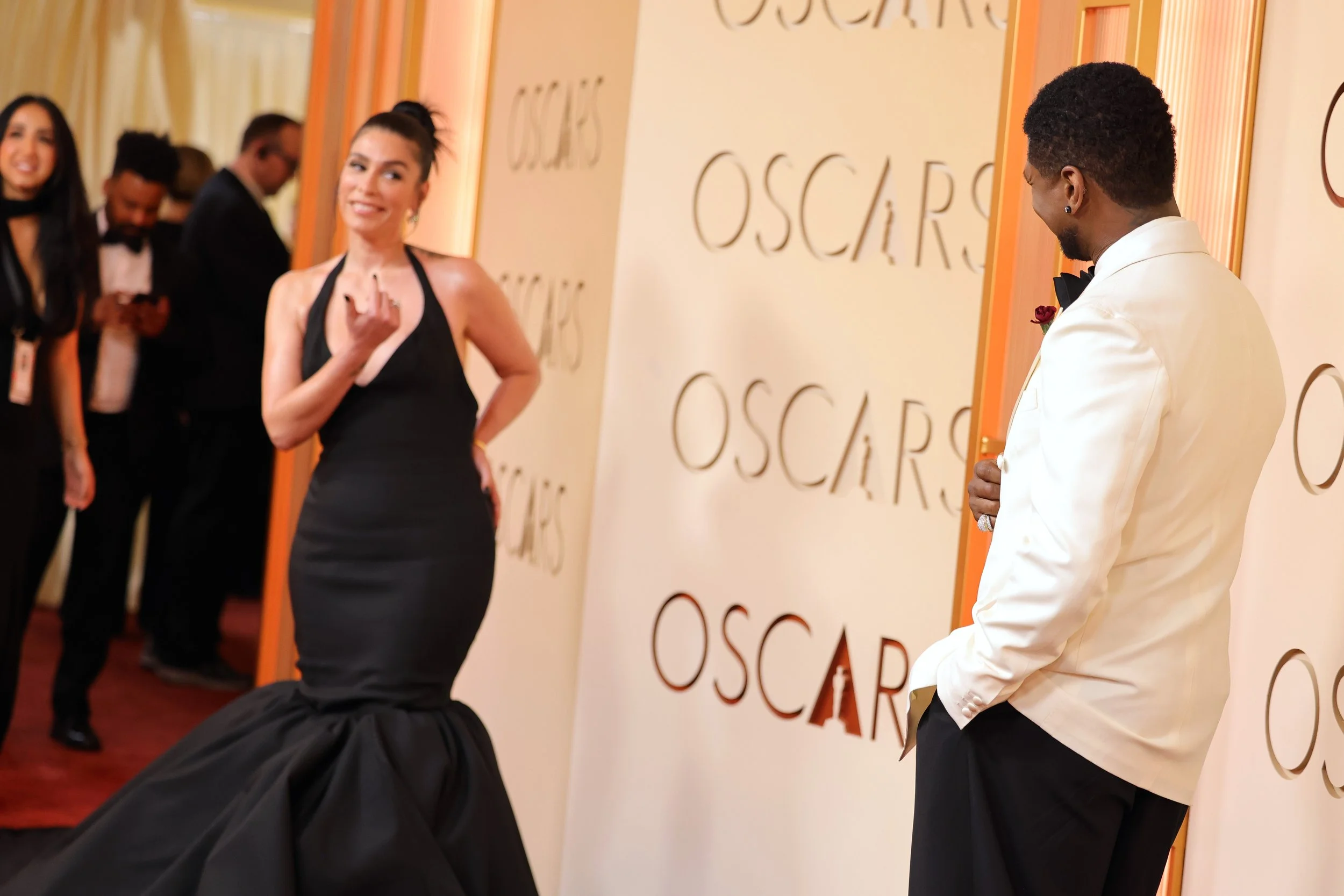 A woman in a black evening gown smiling and posing on the red carpet at the Oscars as a man in a white tuxedo looks on. Other people are visible in the background at the event.