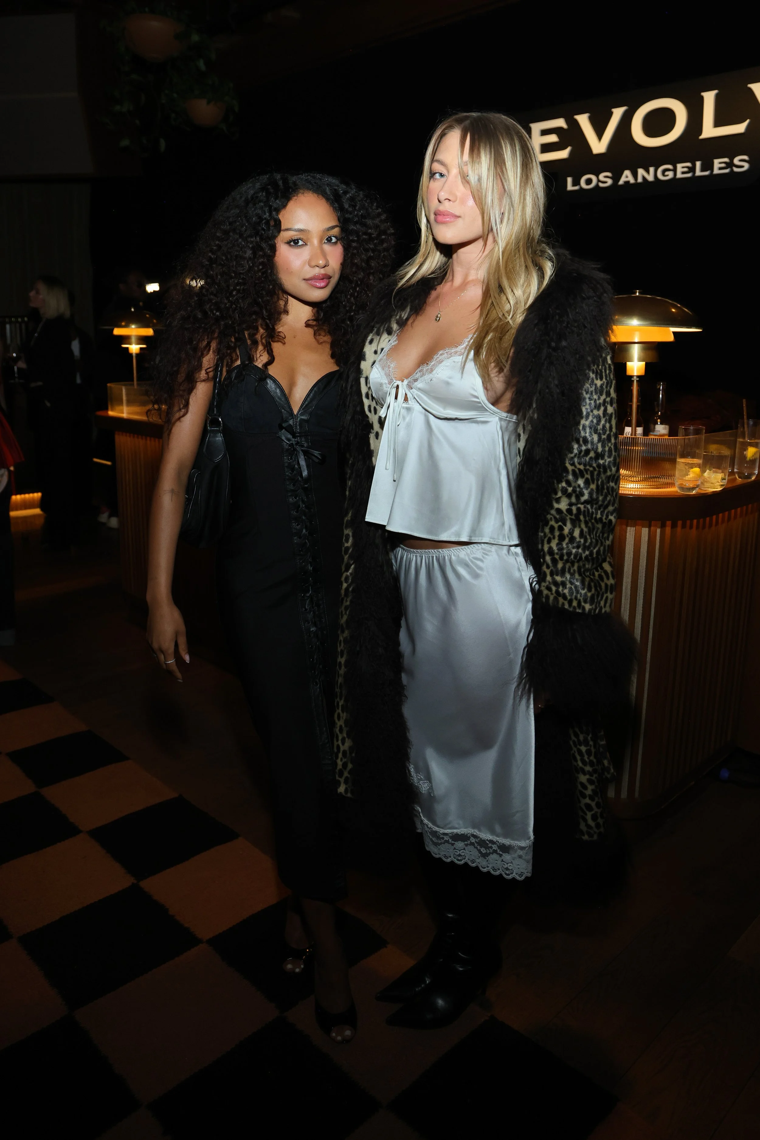 Two women posing inside a dimly lit venue, one with curly hair wearing a black dress, and the other with blonde hair wearing a satin nightgown with a leopard print coat.