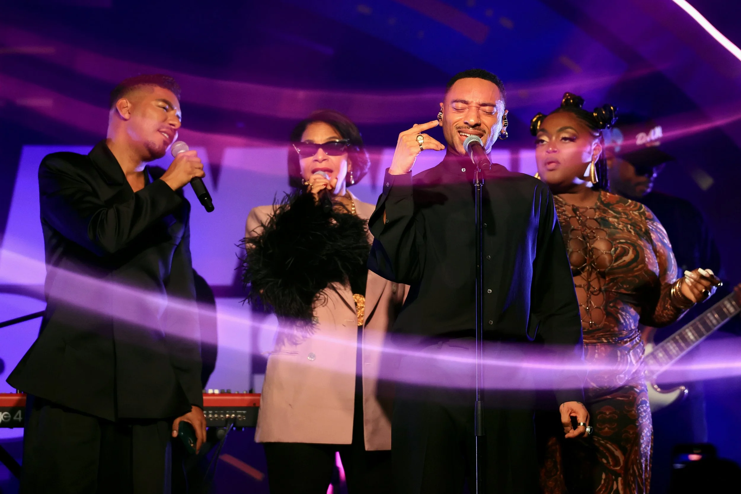 Group of five singers performing on stage with purple lighting and musical equipment.