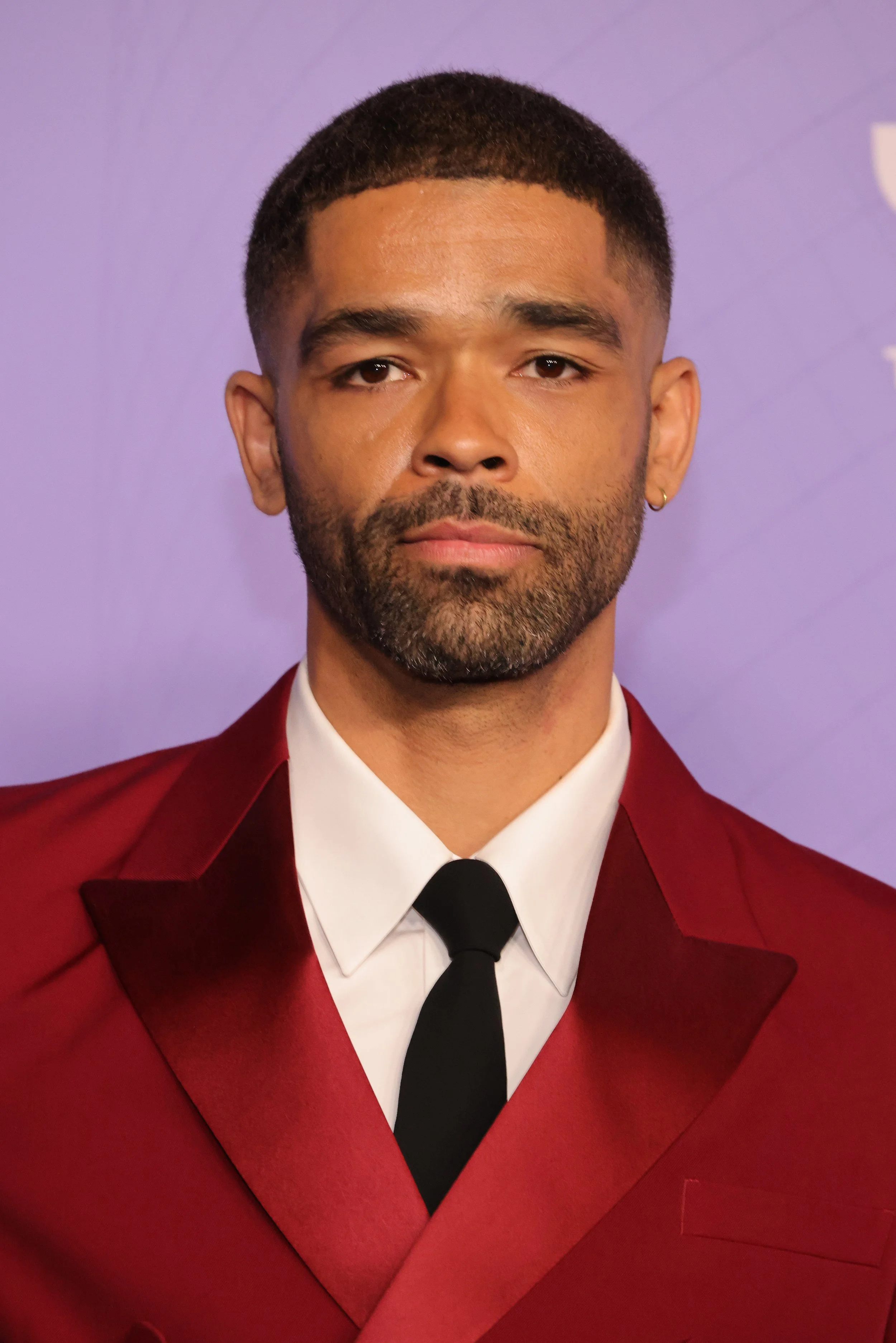 A man with short dark hair, beard, and small earring wearing a red suit with a white shirt and black tie, standing against a purple background.