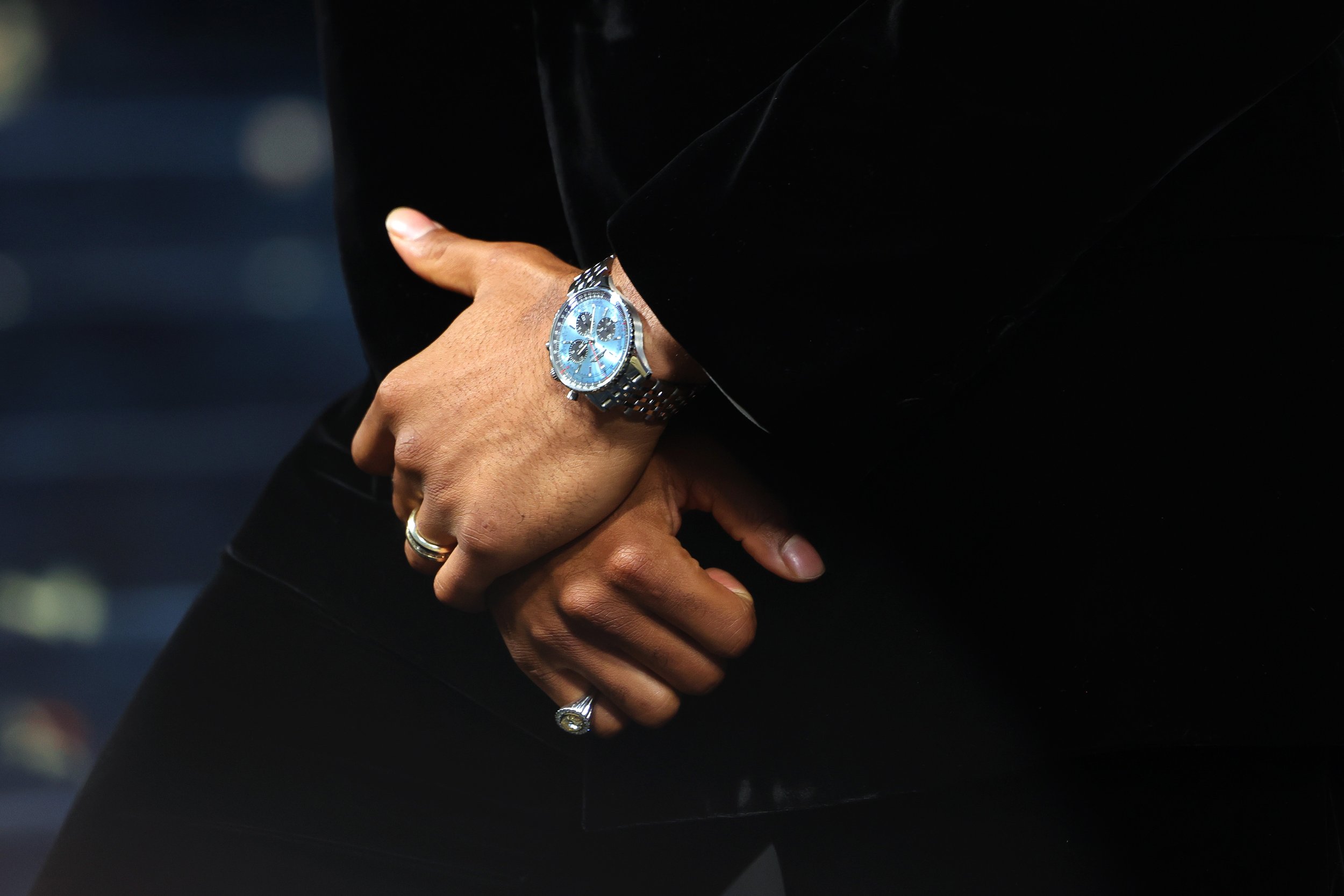 Close-up of a person's hands clasped together, showing a silver wristwatch with a blue face on the left wrist, and several rings, with a dark background.
