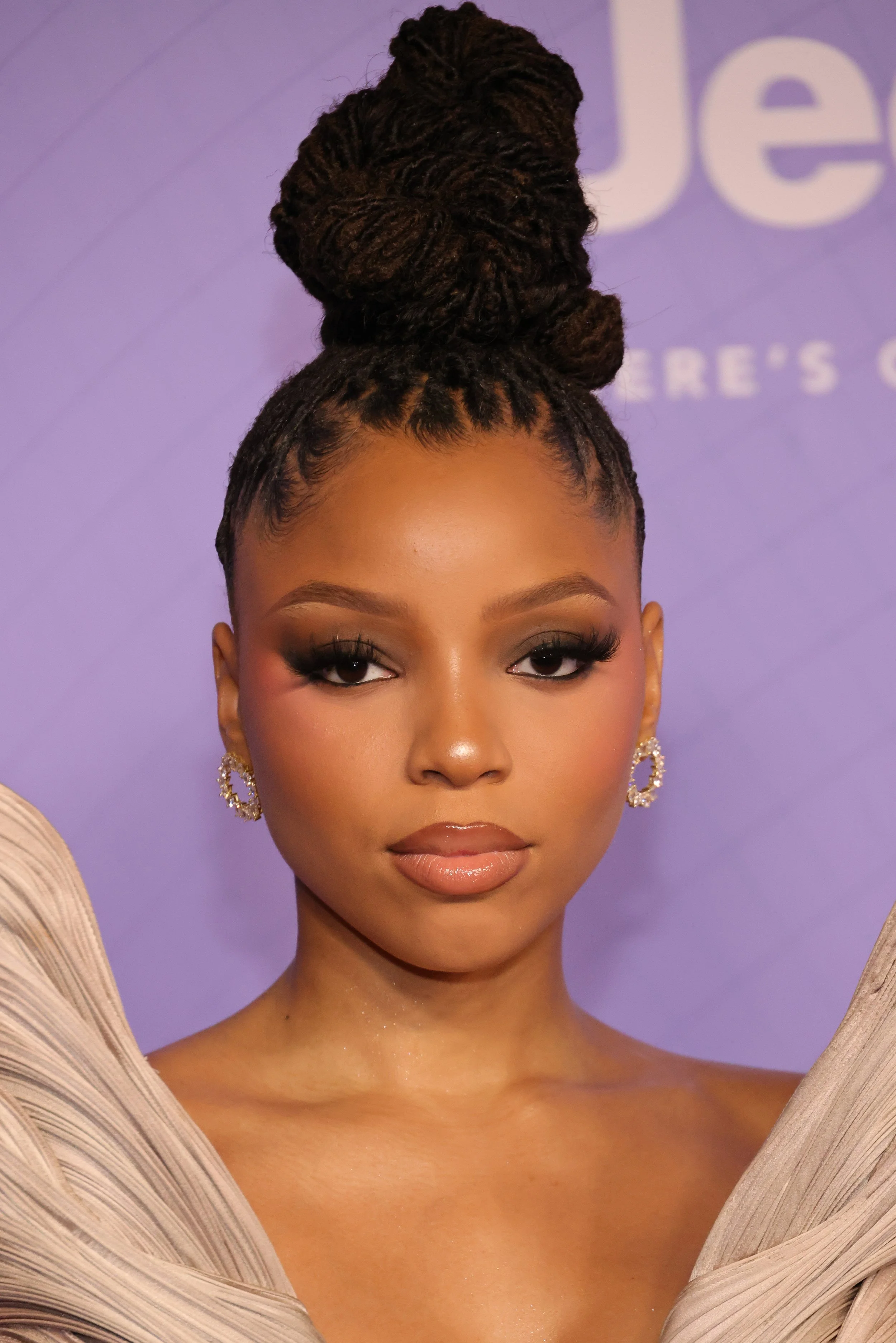 A woman with styled hair in an updo, wearing earrings and makeup, pictured against a purple background.