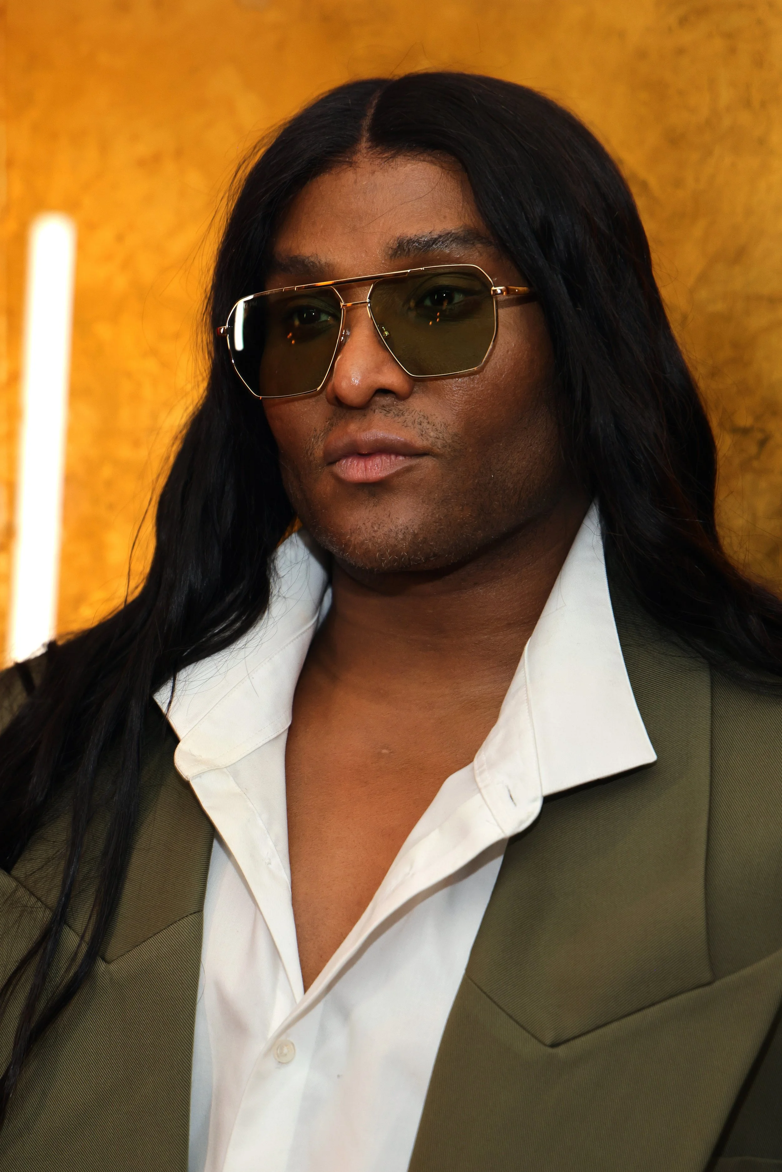A person with long dark hair wearing sunglasses, a white shirt, and a green blazer, standing against a warm-toned background.