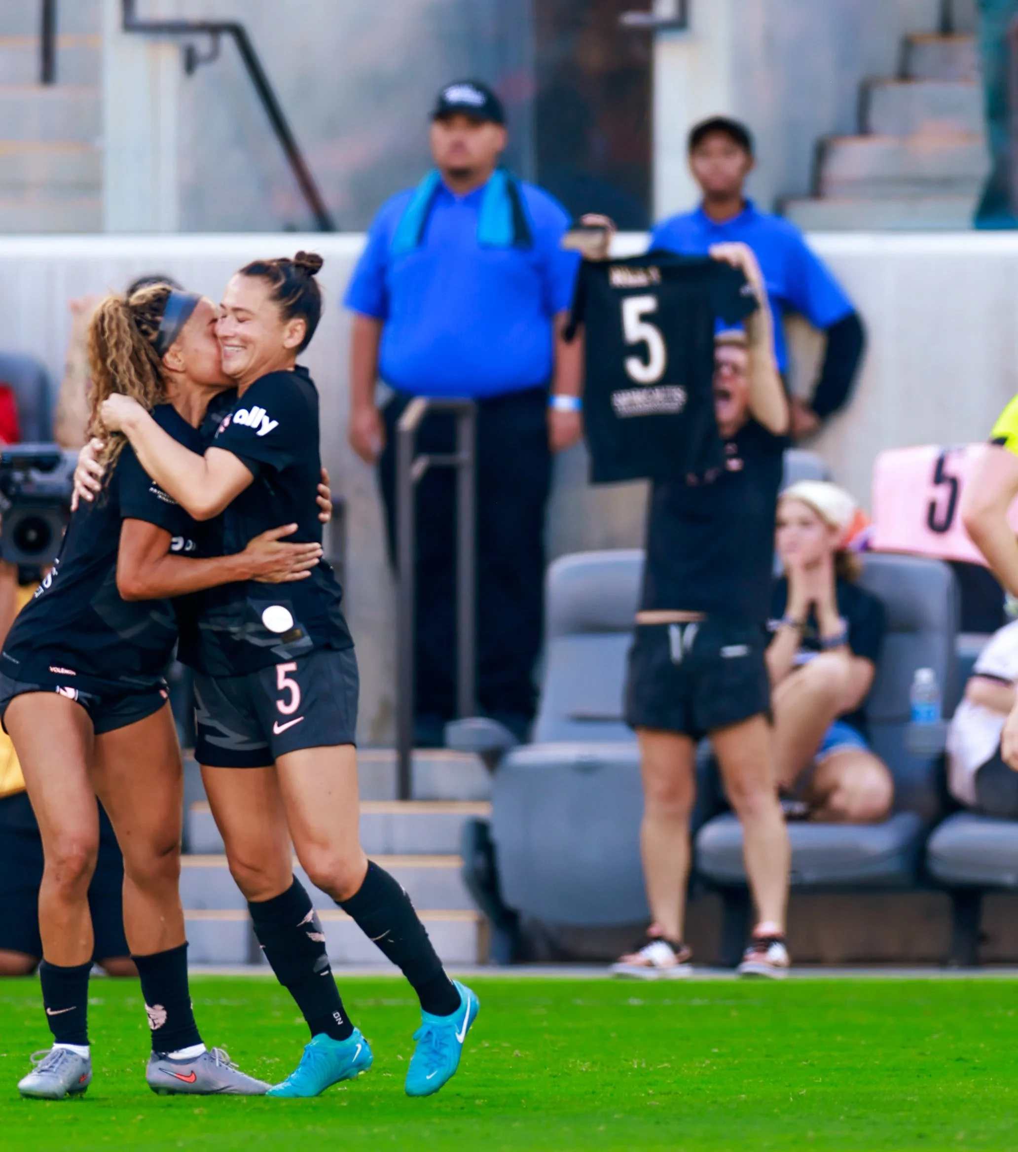 Two female soccer players in black uniforms embracing and celebrating on the field after scoring a goal, with teammates and spectators in the background.