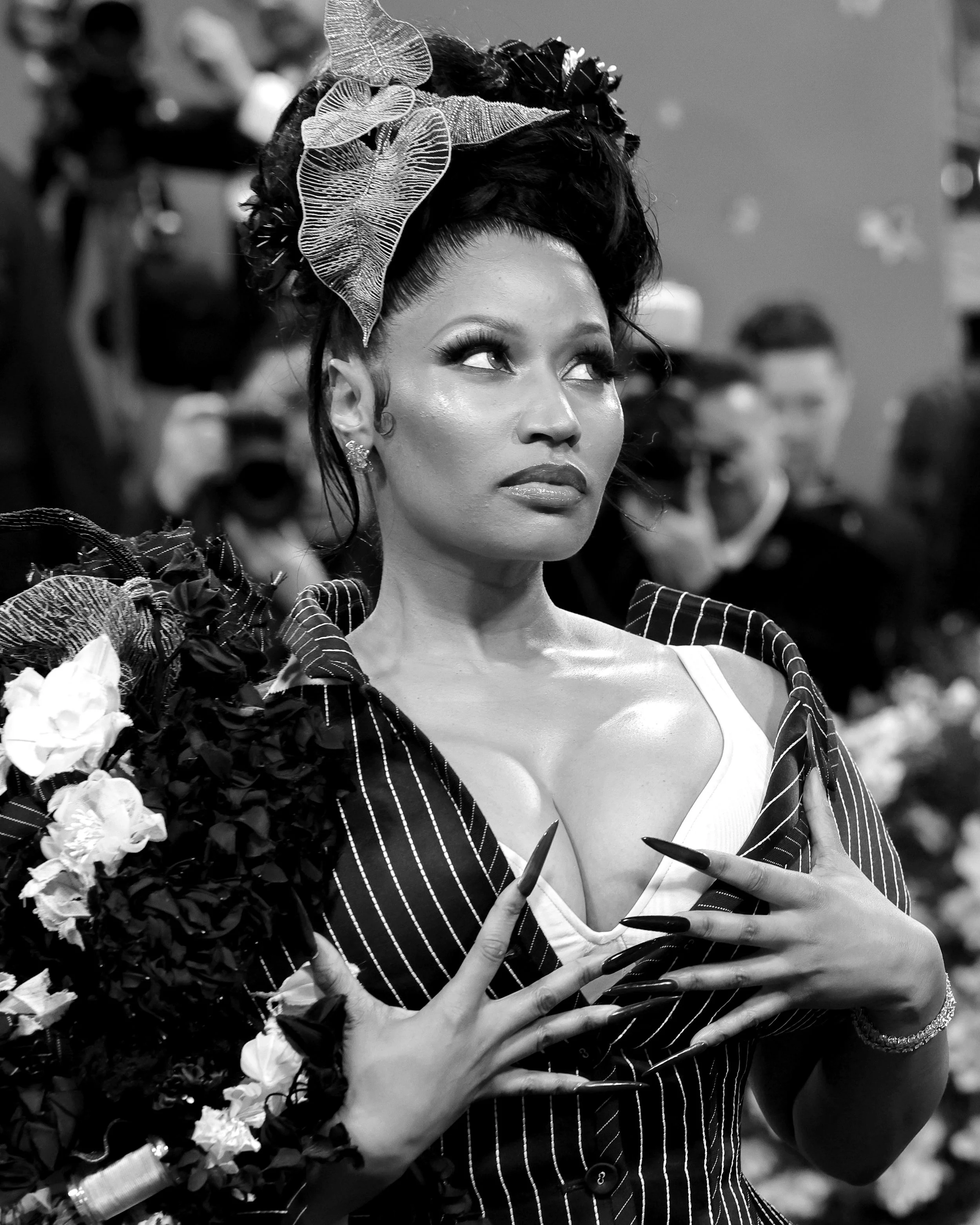 A woman with styled hair and elaborate makeup posing in a fashionable striped outfit, with her hands near her chest, in a black-and-white photo.