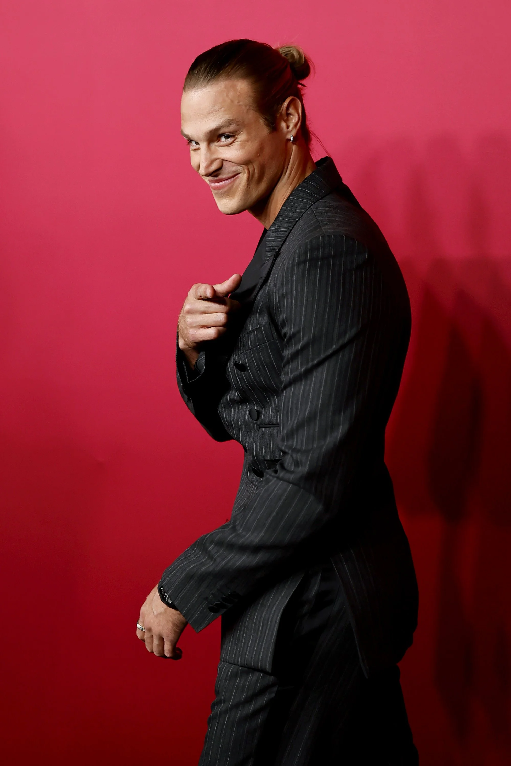 A man with long hair tied in a bun, wearing a dark pinstripe suit, smiling, pointing at himself, standing against a pink background.