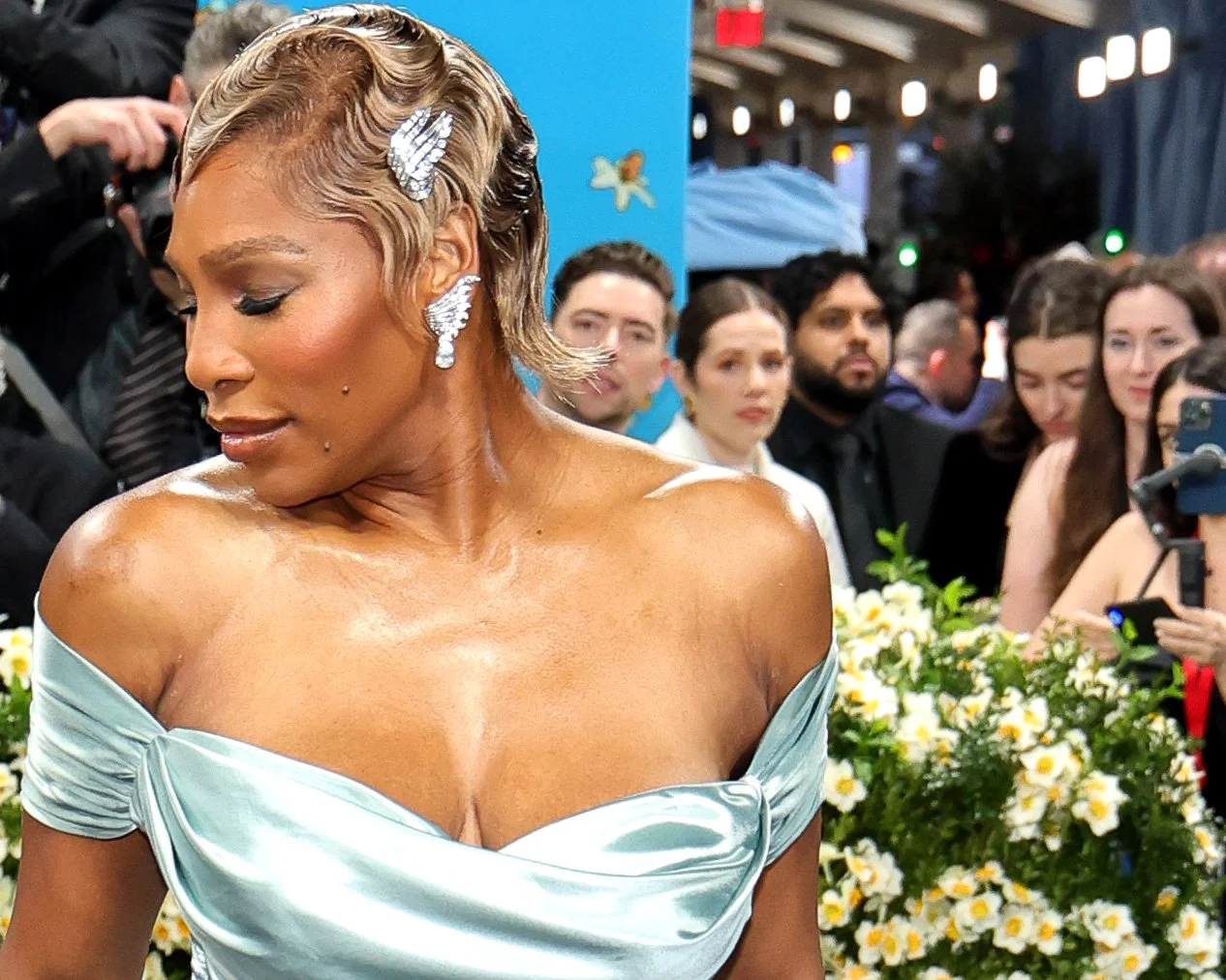 A woman with short, wavy blonde hair wearing large diamond earrings and a light blue, off-shoulder satin dress, surrounded by a crowd of people and a background of flowers at an event.