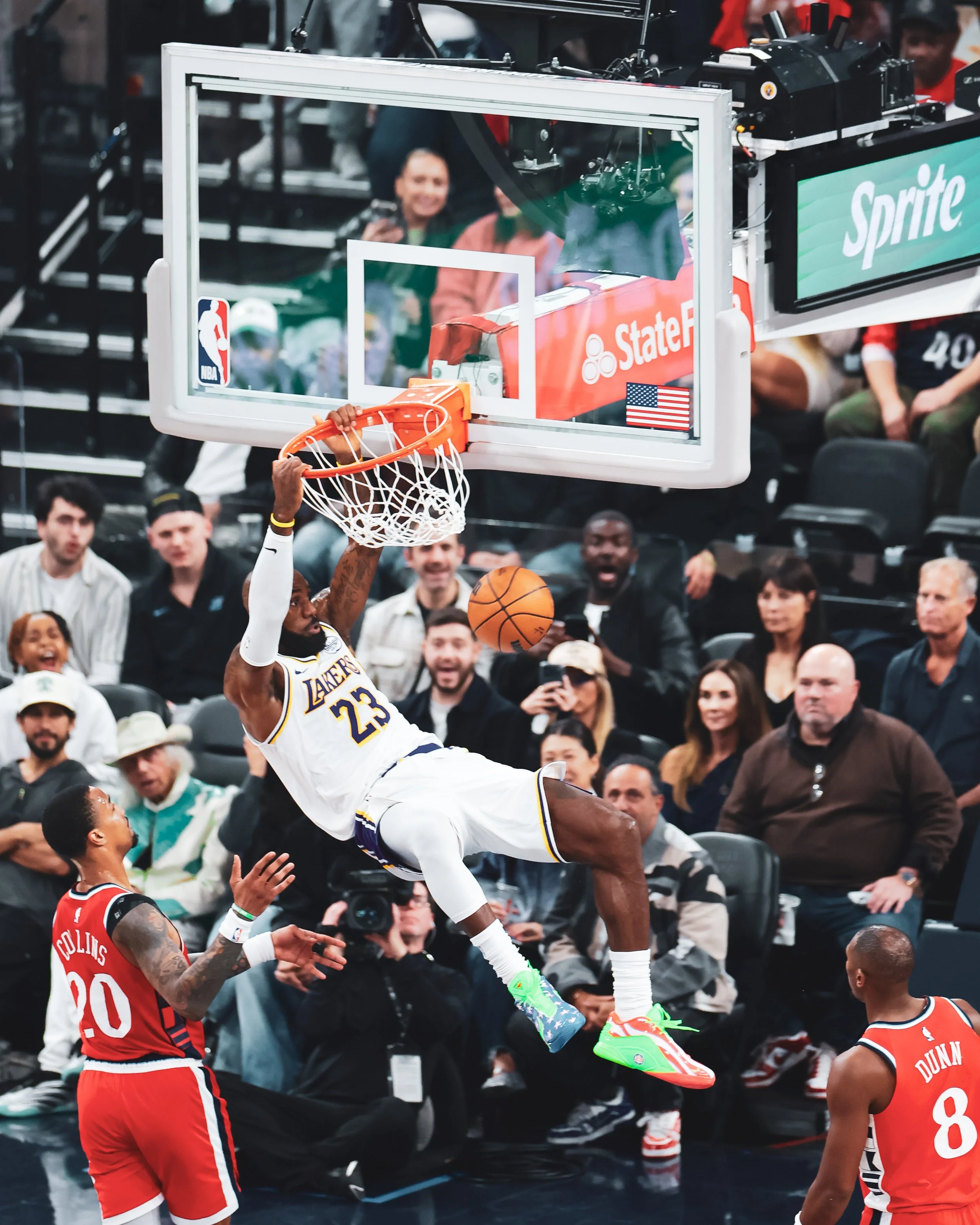 A basketball player from the Lakers, LeBron James, is dunking the ball into the hoop during a game. Two players from the opposing team in red uniforms are nearby, and spectators watch from the stands.