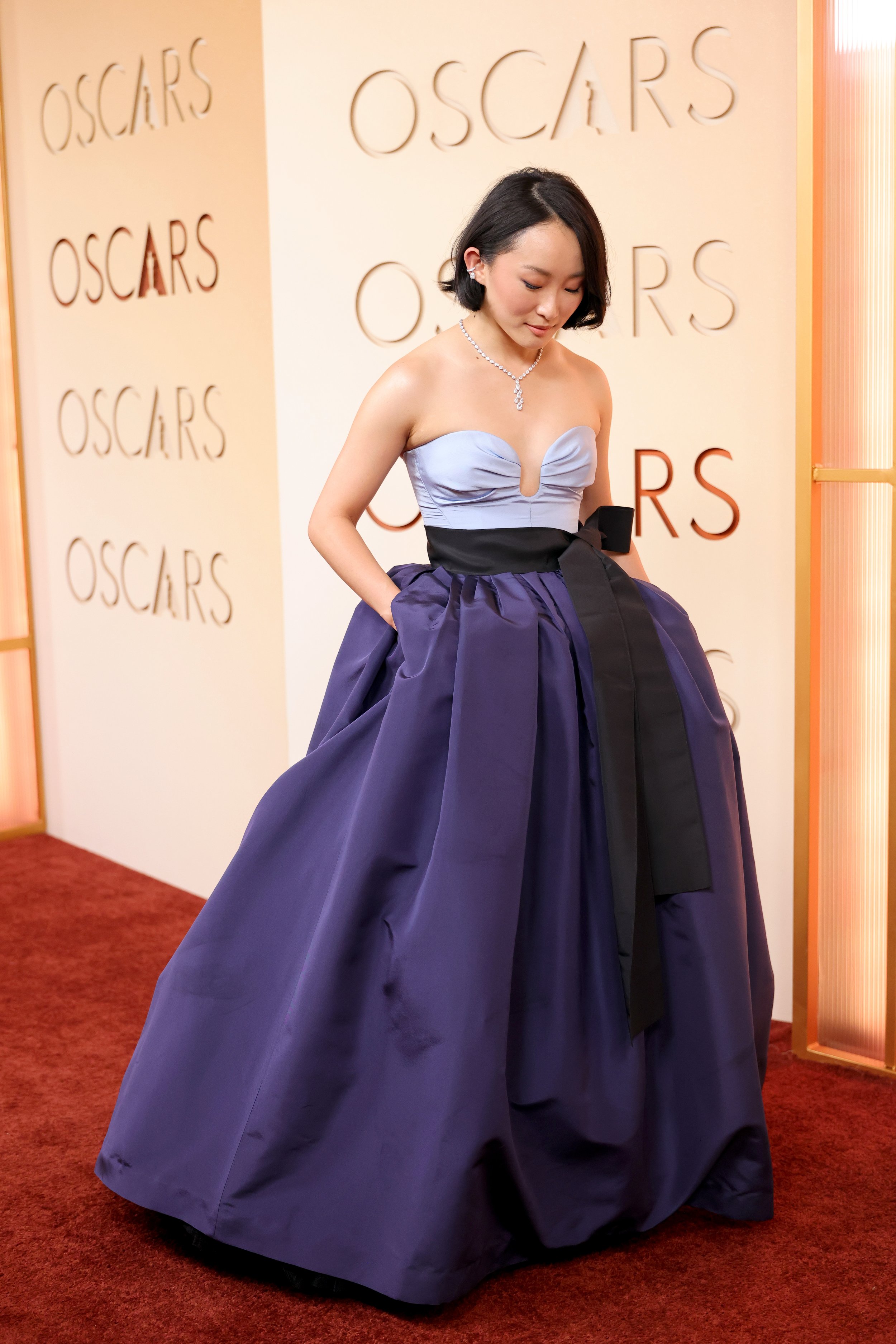 Woman in a strapless purple gown with a black waistband, standing on a red carpet at the Oscars.