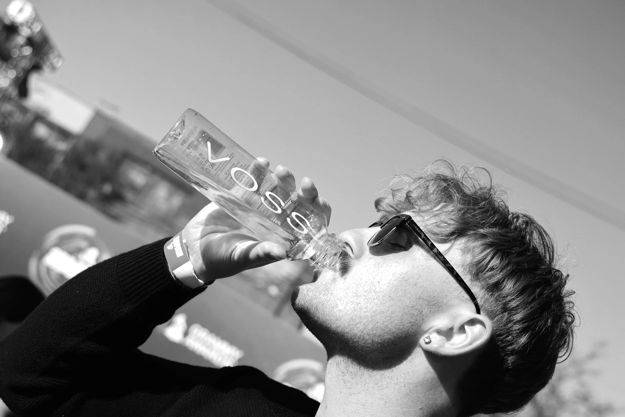 A person with short, curly hair and sunglasses drinks from a large water bottle outdoors, with a clear sky and blurred background.