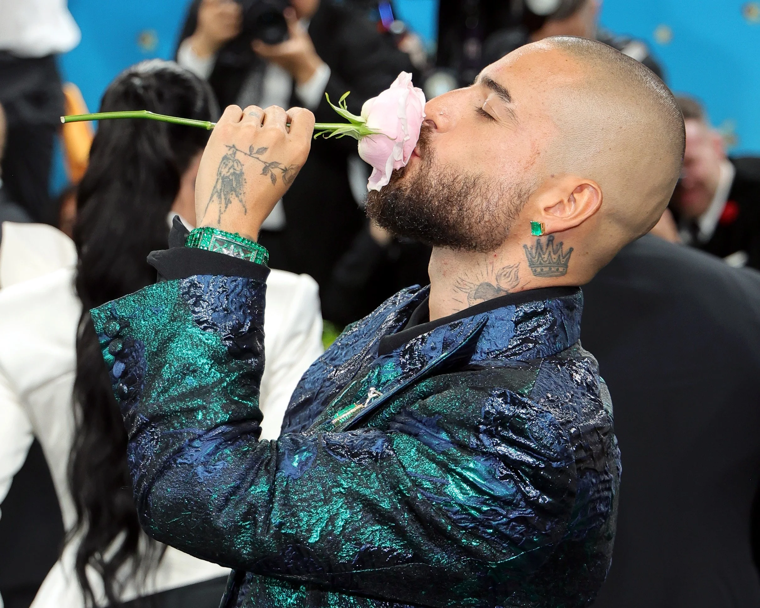 A man with tattoos and a beard is smelling a pink rose, with his eyes closed. He has a shaved head and is wearing a vibrant, patterned jacket. There are people in the background, some holding cameras, in an indoor setting.
