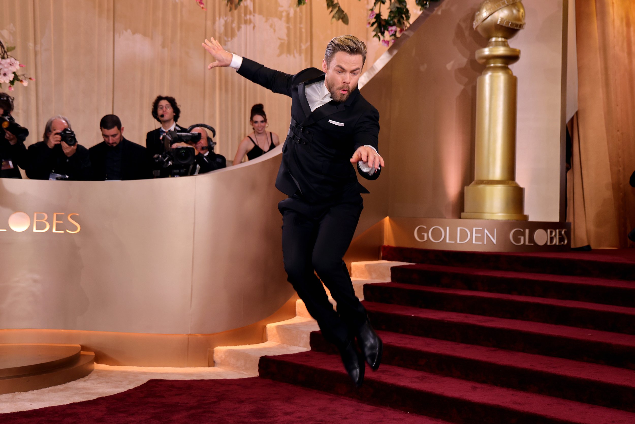 A man in a tuxedo appears to be dancing or jumping on red carpet stairs at the Golden Globes awards event. There are photographers and onlookers behind him, and a large Golden Globes statue is visible in the background.