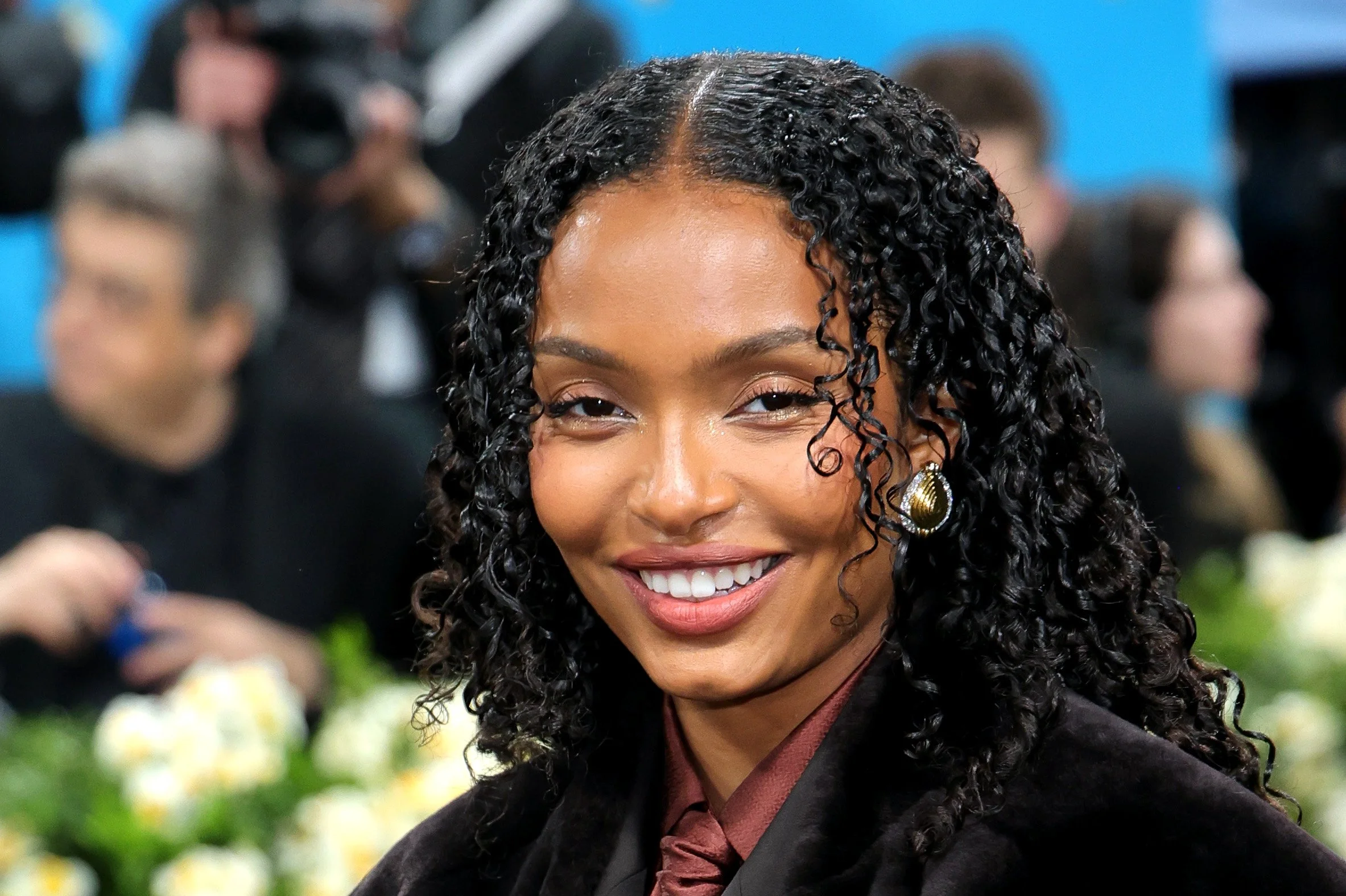 A woman with curly black hair smiling at an event, wearing a black blazer and gold earrings, with people and floral arrangements in the background.