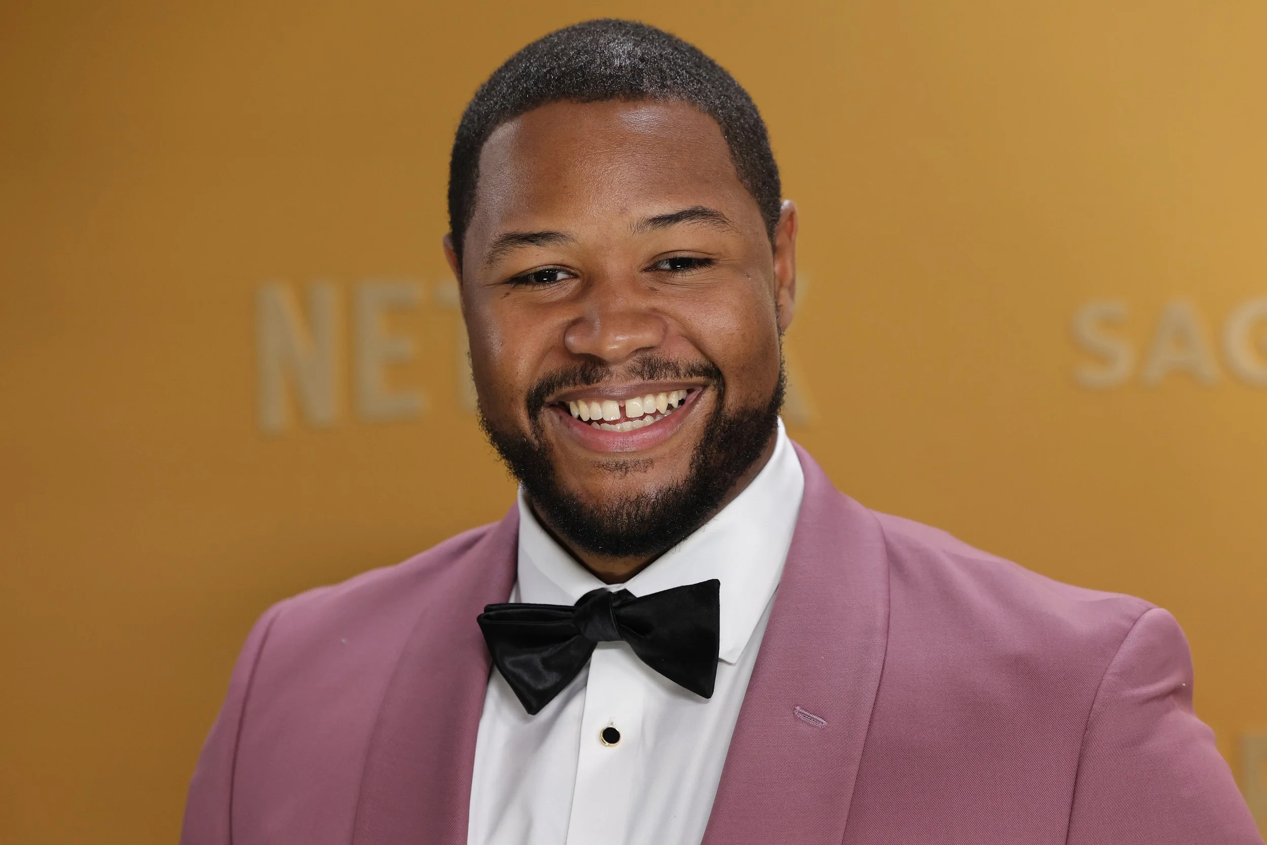 A smiling man with a beard wearing a pink blazer, white shirt, and black bow tie, standing against a yellow background.