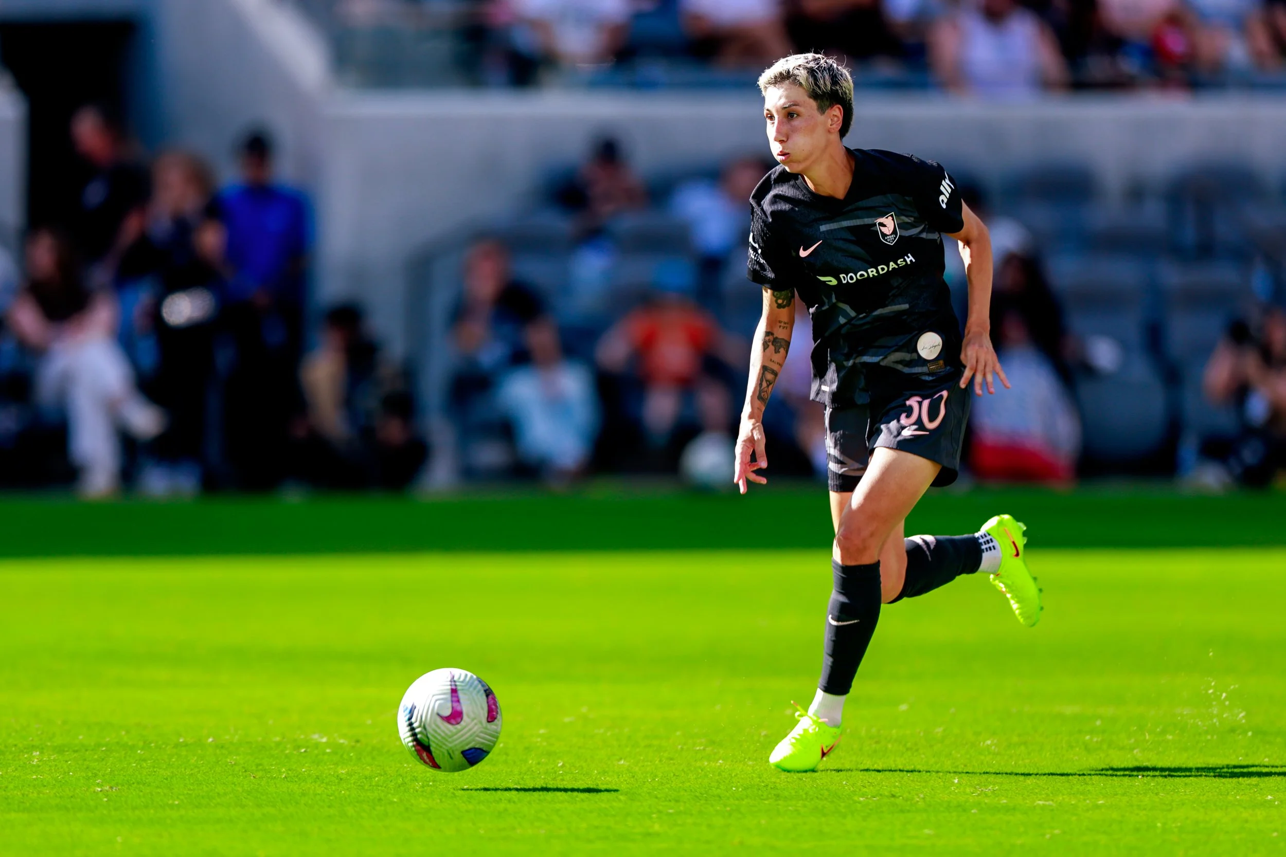 A soccer player is running on a green field with a soccer ball in front of him. The player is wearing a black jersey, black shorts, and bright yellow-green soccer cleats. Fans can be seen in the background sitting on bleachers.