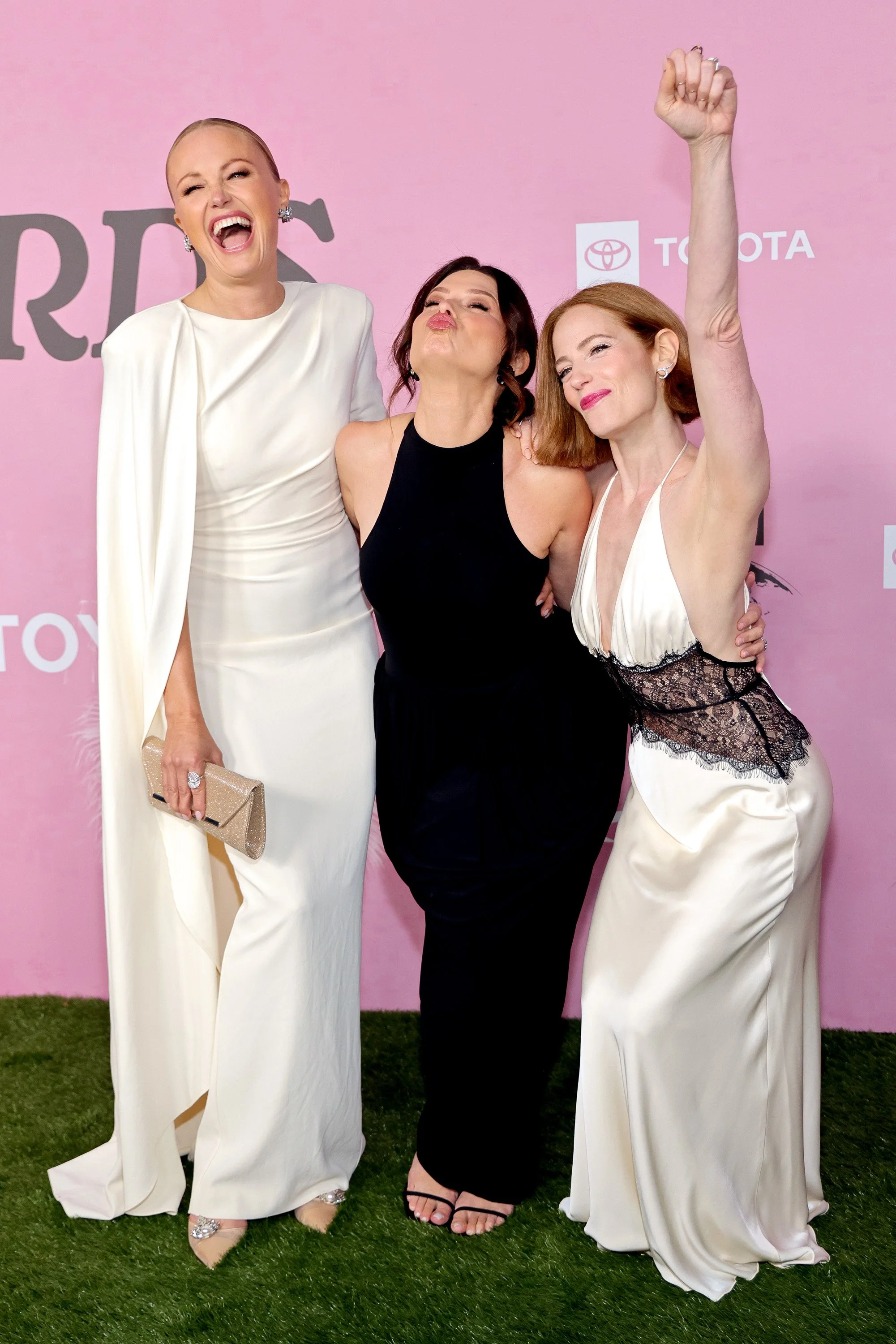 Three women dressed in elegant gowns celebrating together at an event with a pink backdrop.