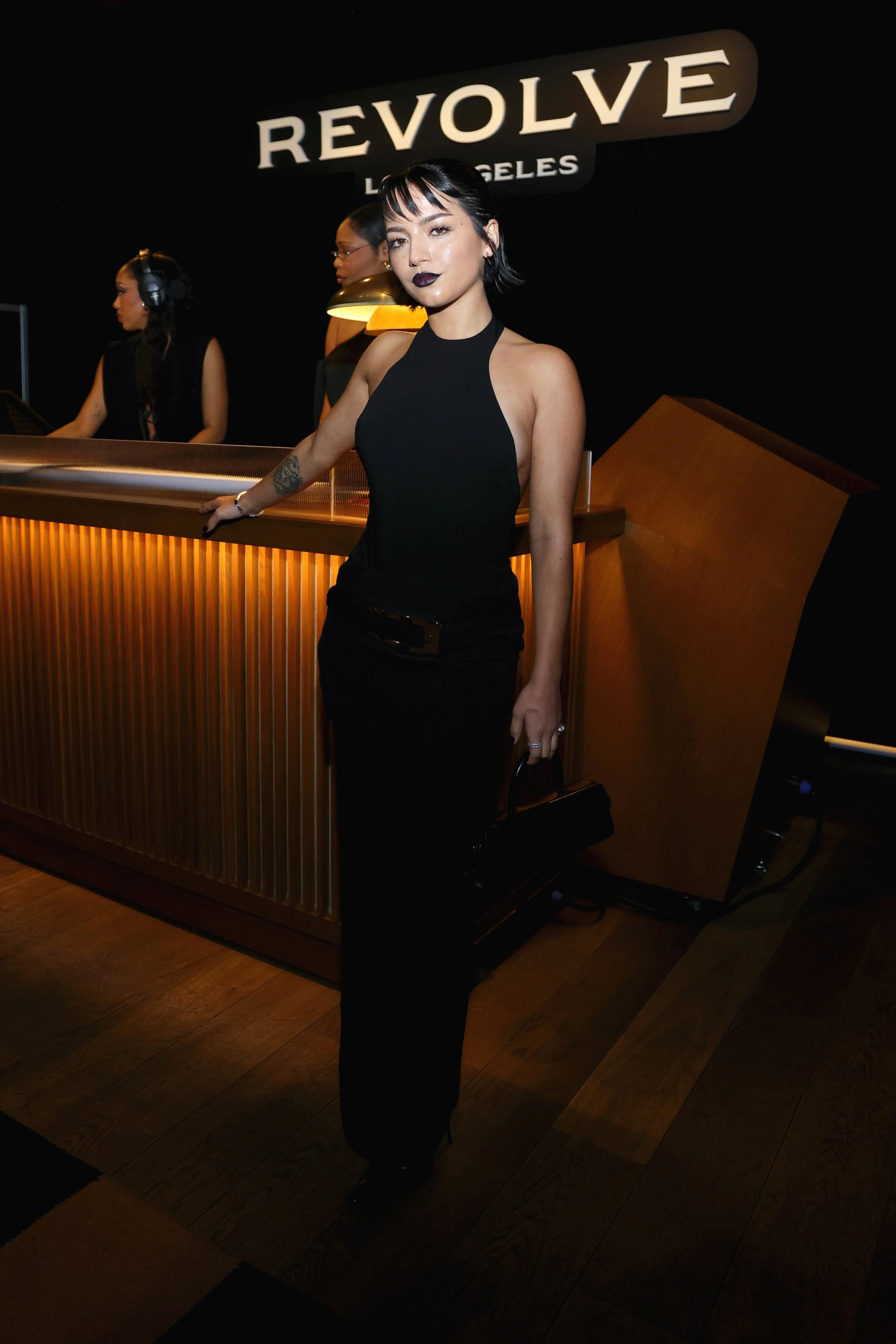 A woman in a black sleeveless dress standing at a wooden reception desk inside a nightclub or bar, with a sign that reads 'REVOLVE LOS ANGELES' in the background.