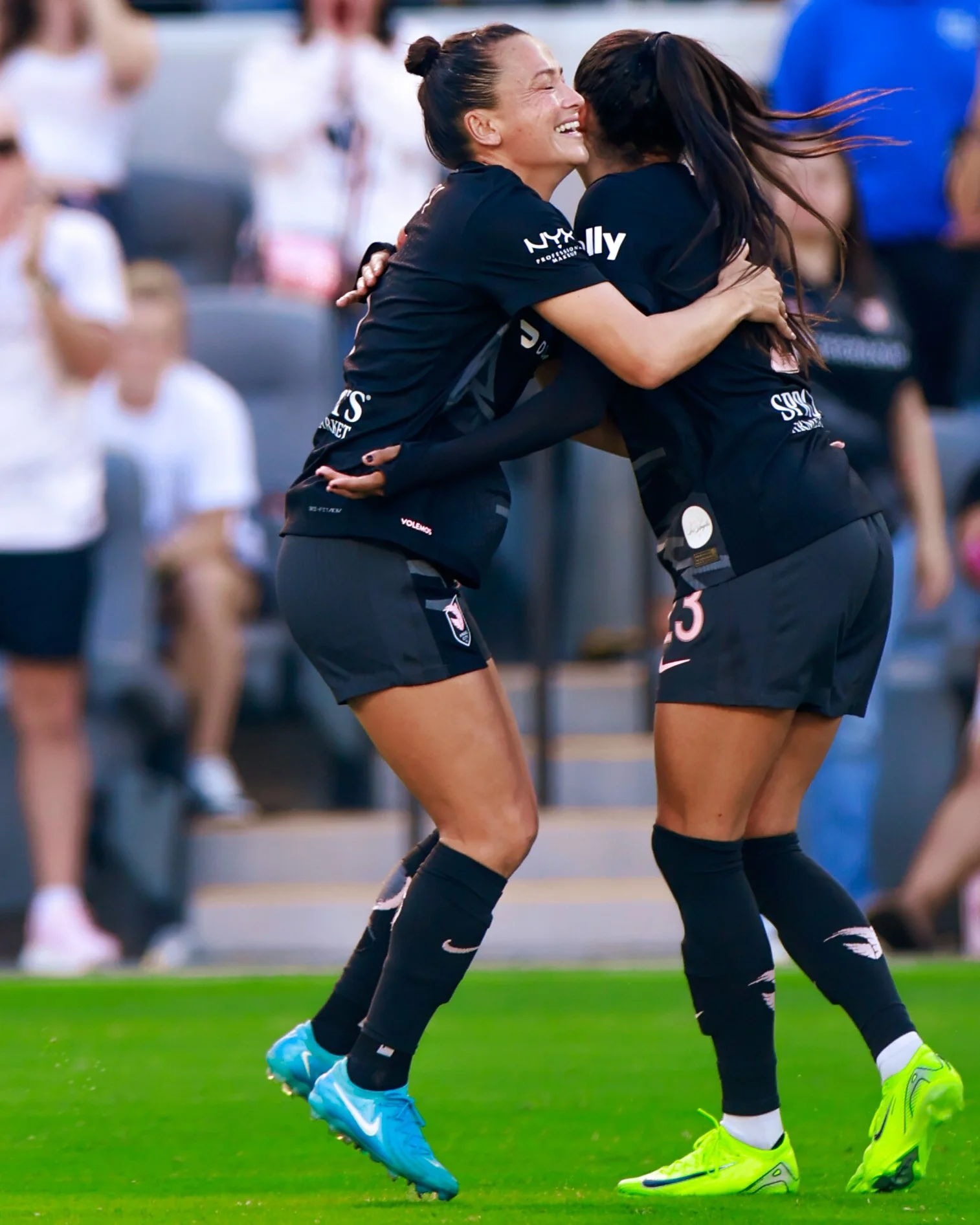 Two female soccer players in black uniforms hugging and celebrating on the field.