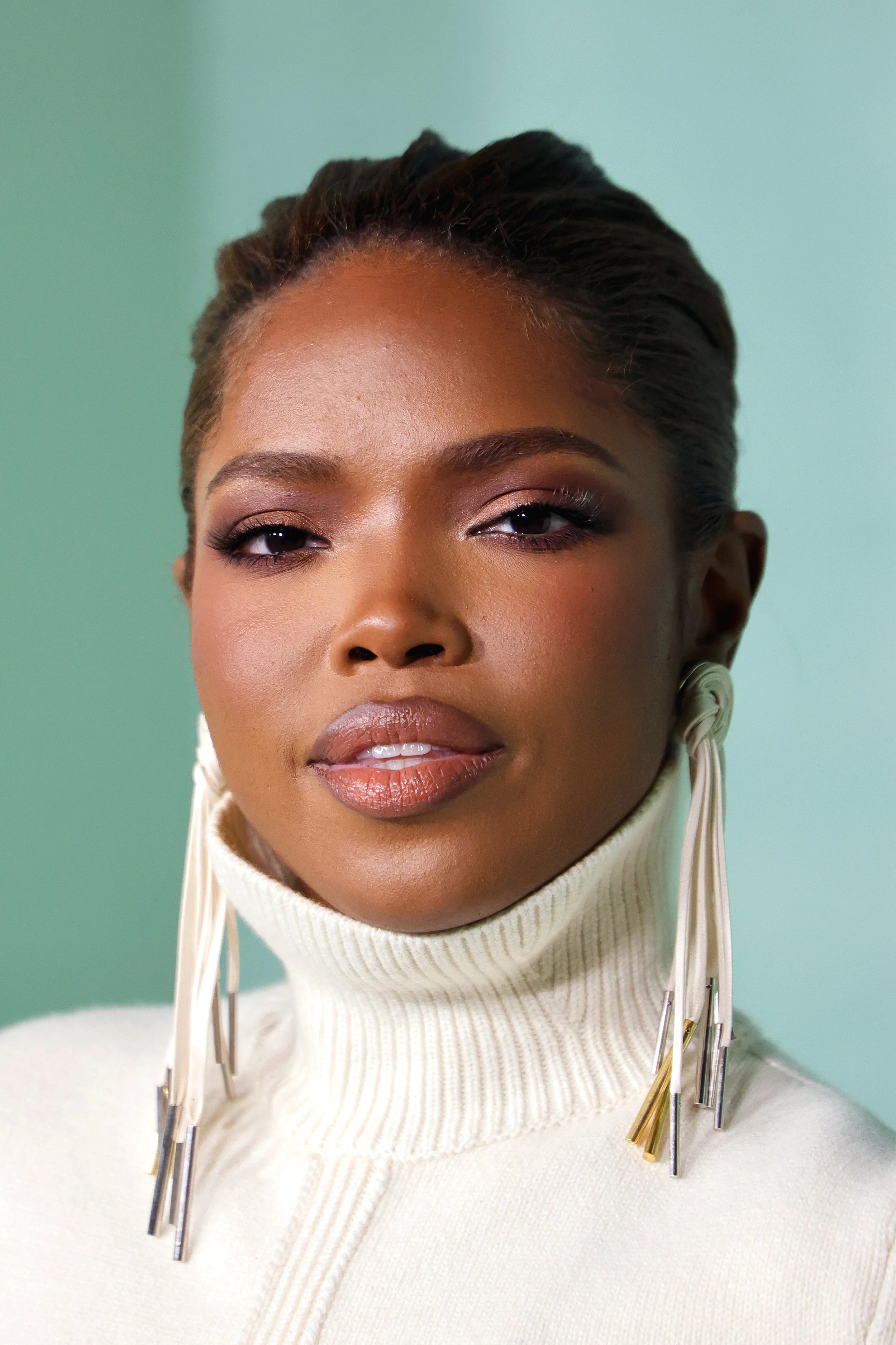 Close-up of a woman with styled hair, wearing a white turtleneck sweater and statement earrings, with neutral makeup against a pale green background.