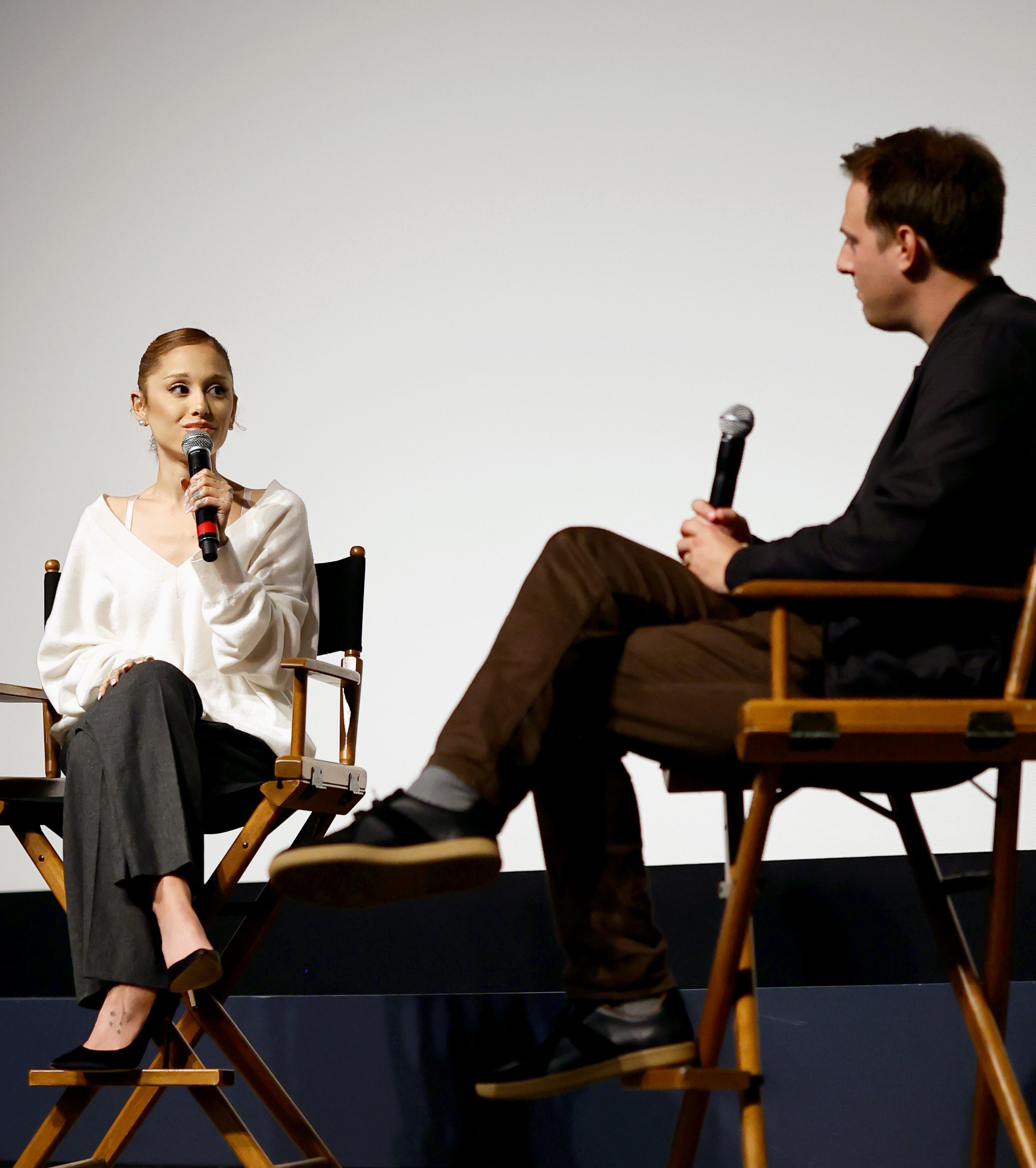 A woman and a man sitting on stage in director's chairs, having a conversation during a talk or interview. The woman is holding a microphone and looking at the man, who is also holding a microphone.