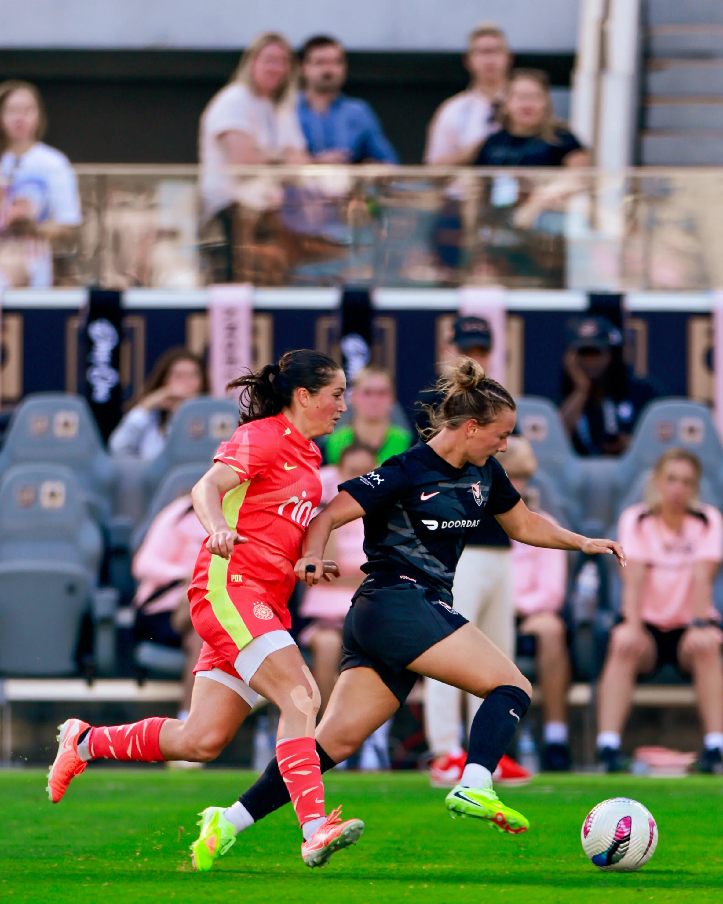Two female soccer players running after the ball during a match, with spectators watching from the sidelines.